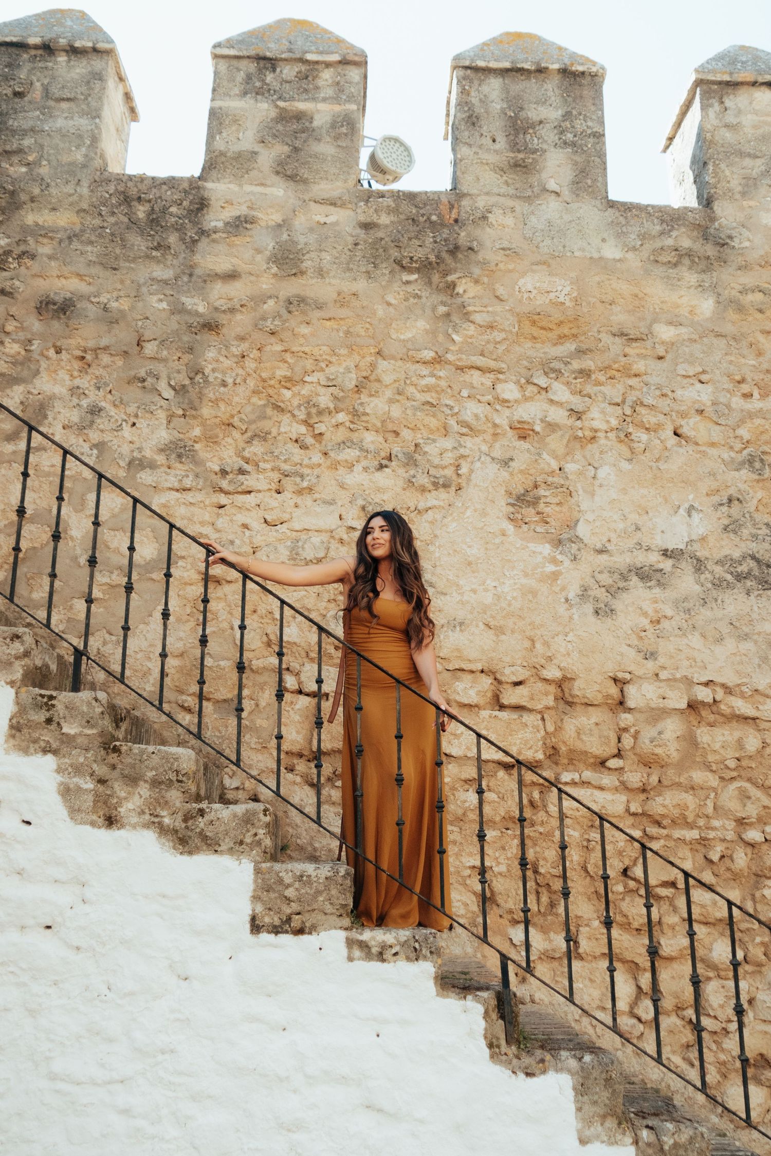 Woman in brown maxi dress posing on ancient stone castle stairs with iron railing.