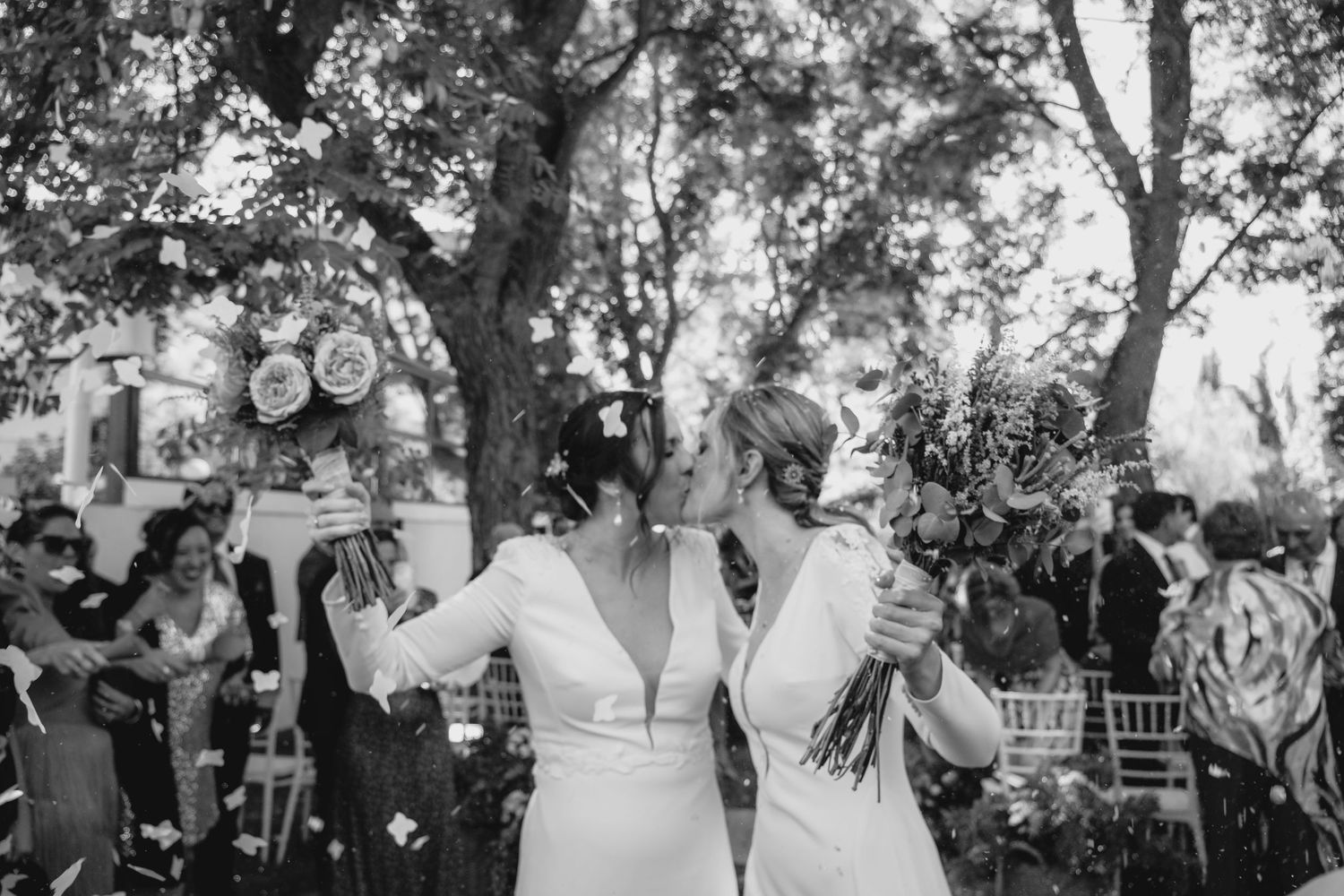Two brides in white gowns share a kiss while guests celebrate with confetti at an outdoor wedding ceremony.
