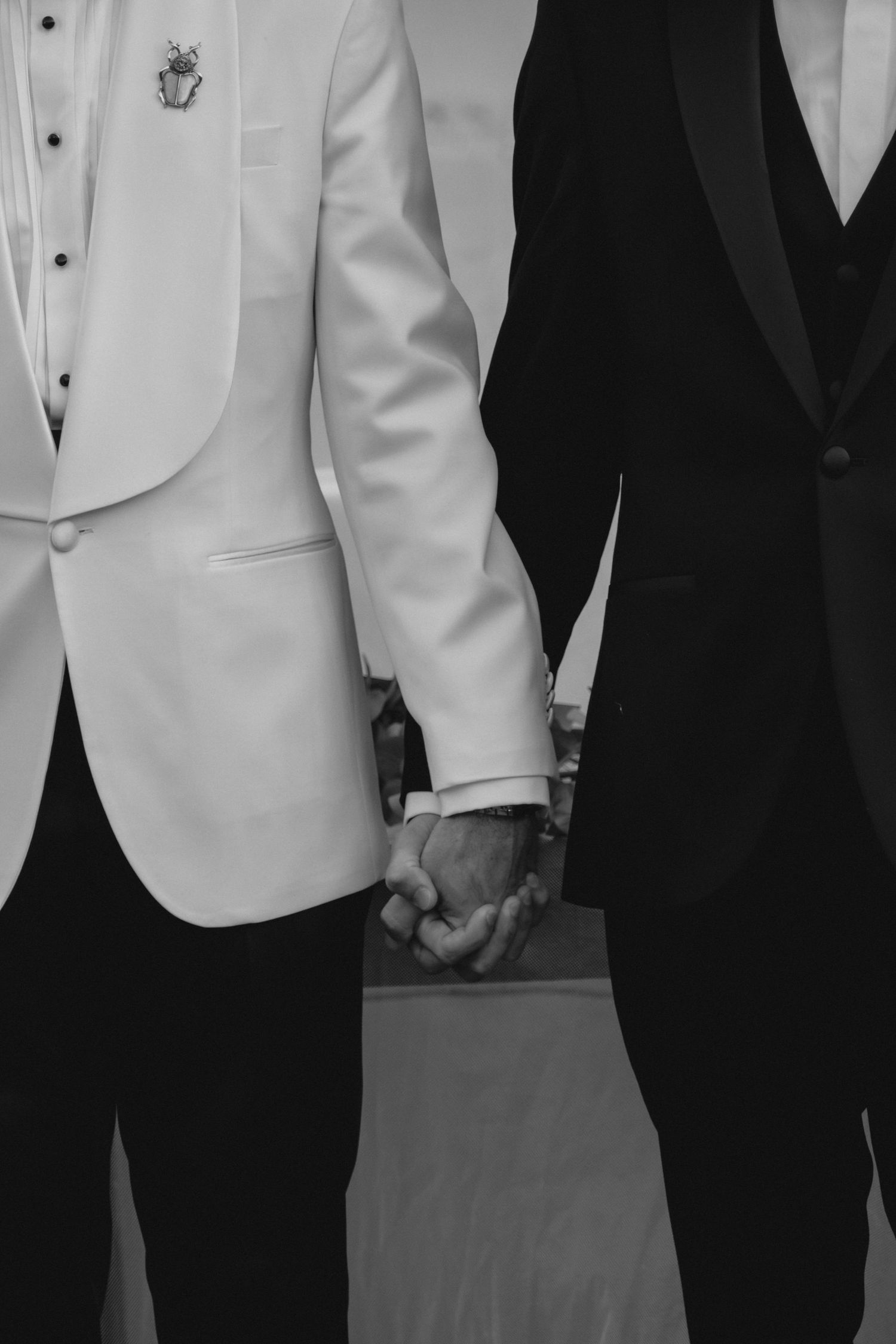 Black and white close-up of two grooms holding hands in white and black tuxedos at wedding.
