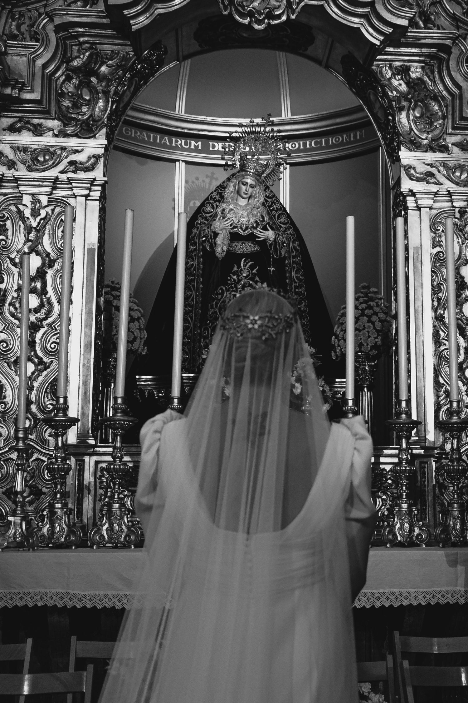 Bride in white gown and veil praying before ornate baroque altar in black and white.