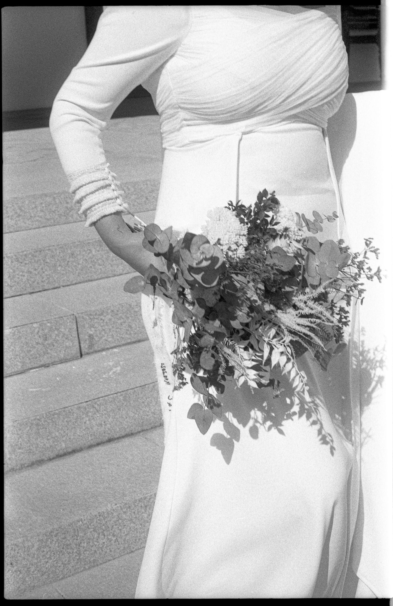 Bride in white long-sleeve gown holding lush greenery bouquet on stone steps, black and white photo.