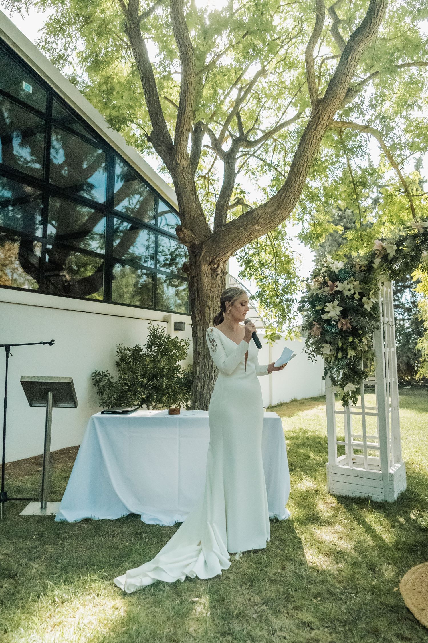 Bride in long white gown speaks at outdoor wedding ceremony beside floral arch and tree.