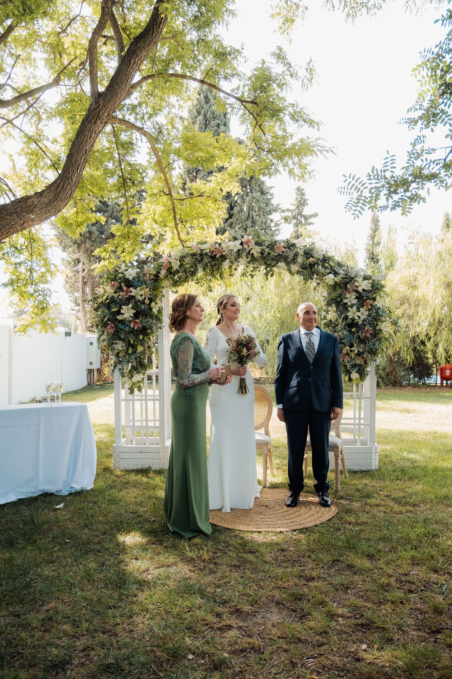 Bride stands with officiant and guest under floral arch at outdoor garden wedding.