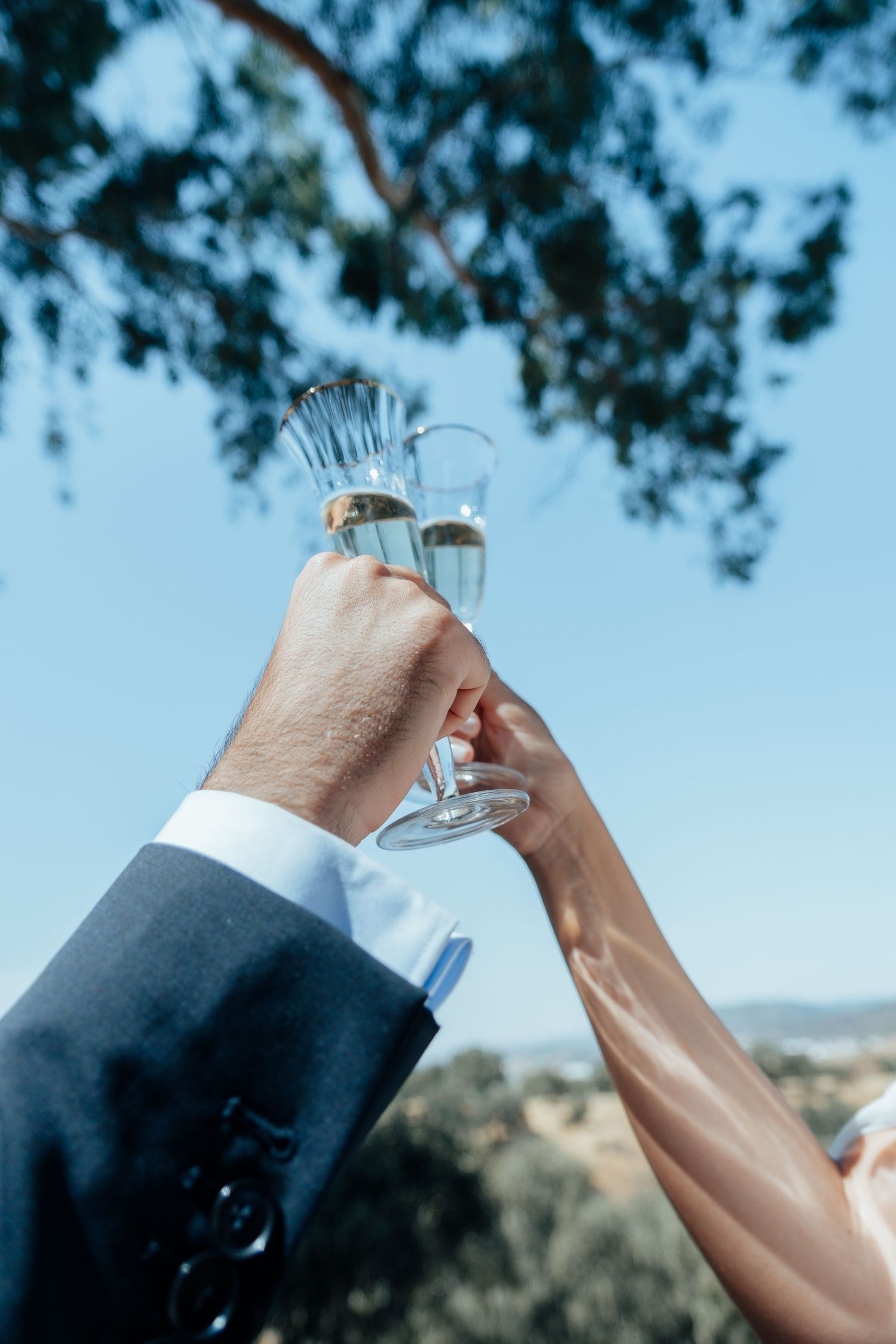Bride and groom toasting with crystal champagne flutes against a clear blue sky at an outdoor wedding celebration.