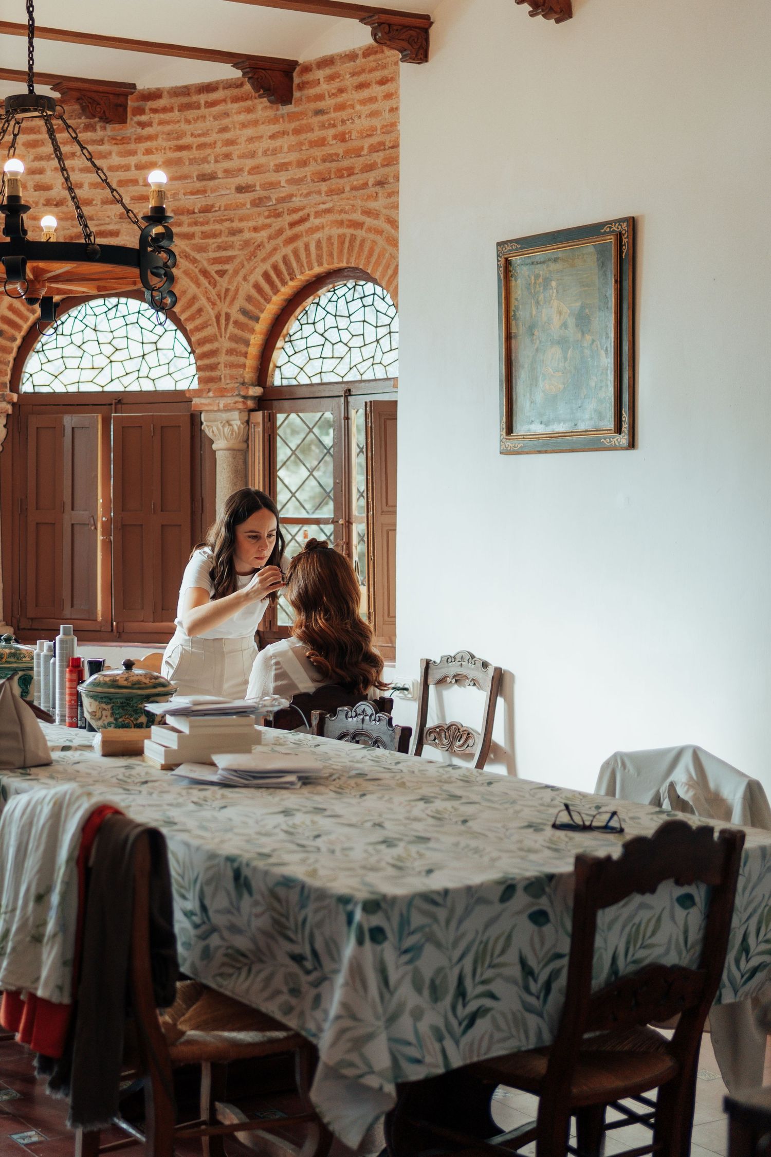 Woman styling another womans hair at a rustic dining table in an elegant historic venue.