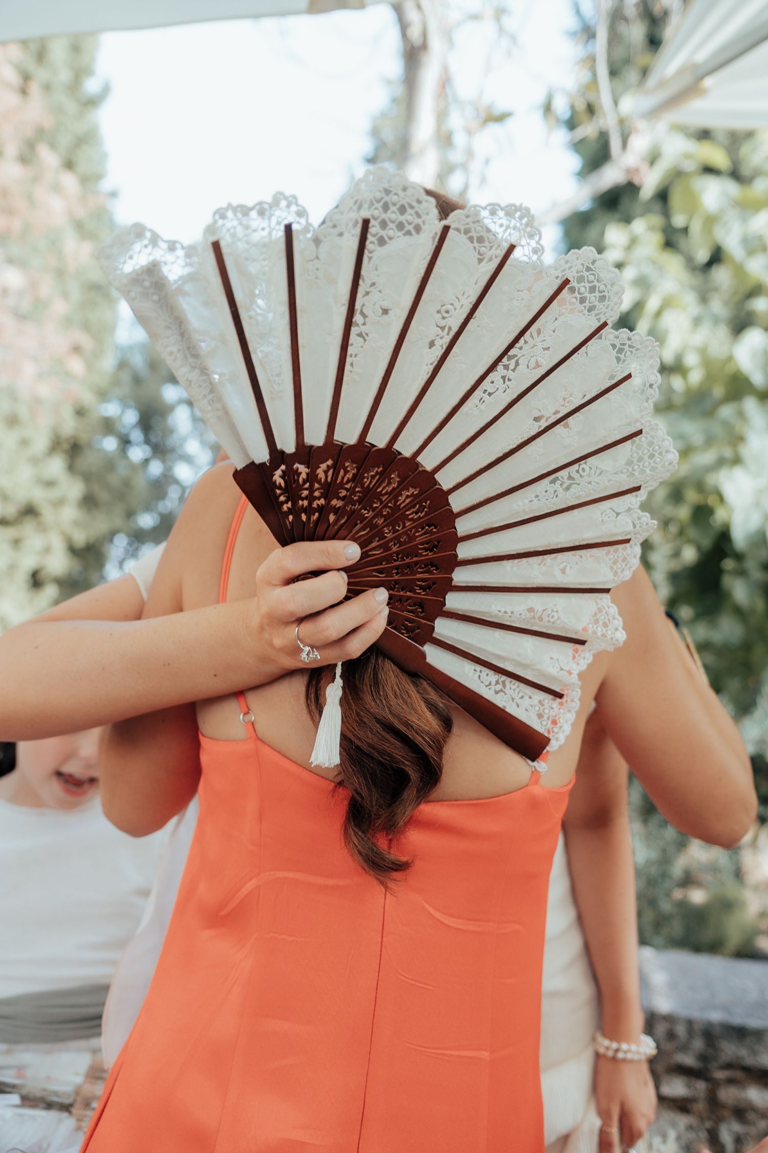 Woman in orange dress holding decorative white lace hand fan at outdoor wedding celebration.