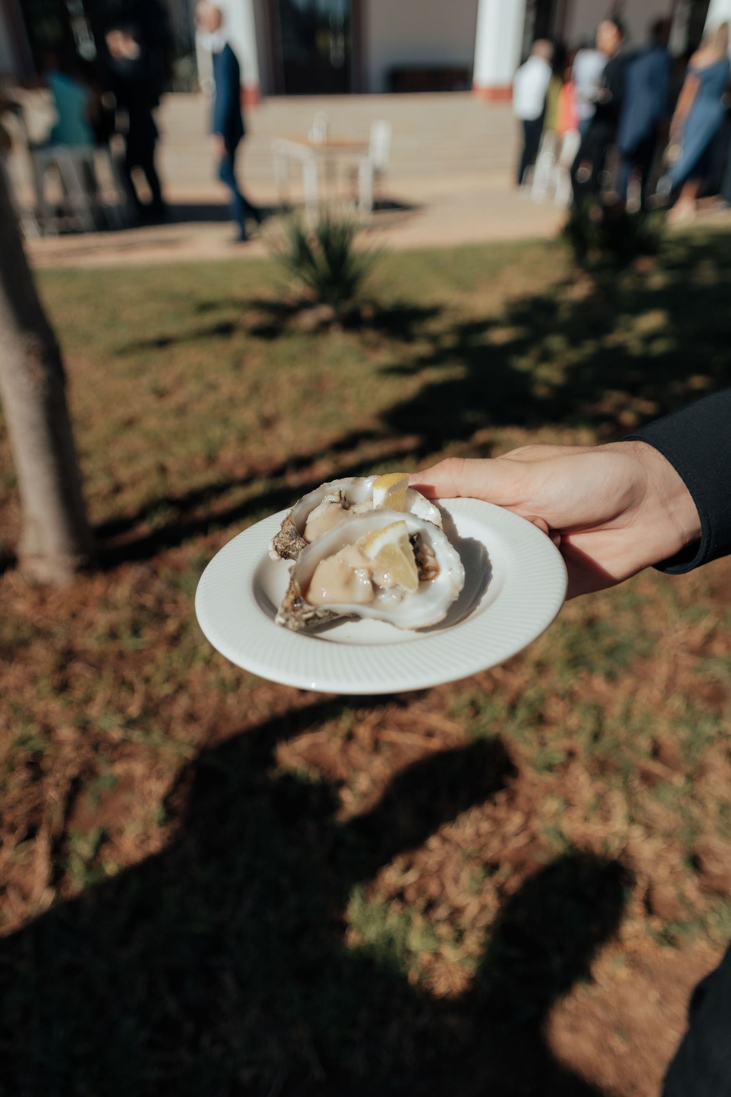 Hand holding a white plate with a fresh oyster at an outdoor wedding reception.