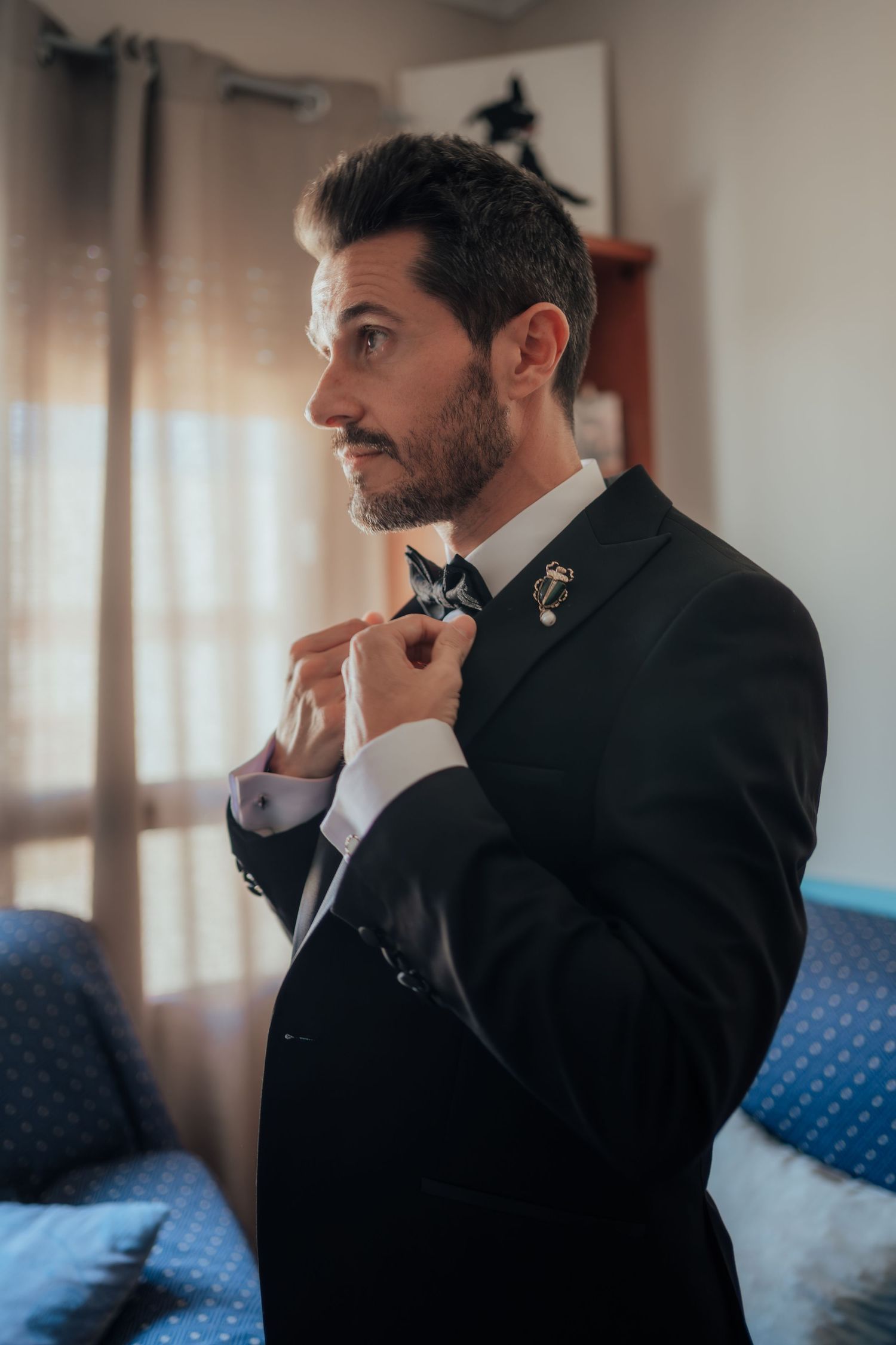 A bearded groom in a black suit with bow tie and beetle brooch adjusts his collar in profile view.