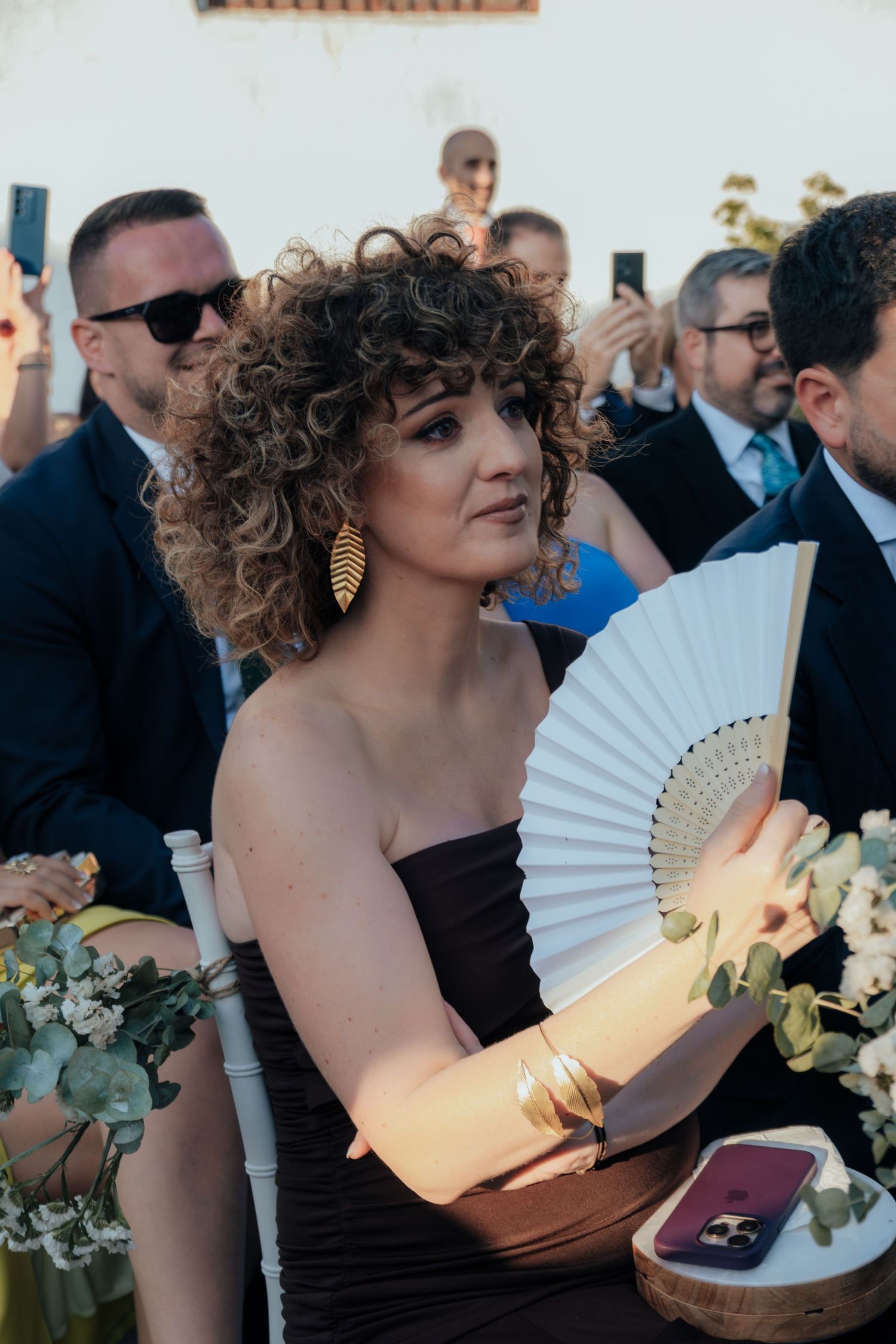 Wedding guest with curly hair holds white fan wearing strapless brown dress and gold leaf earrings.