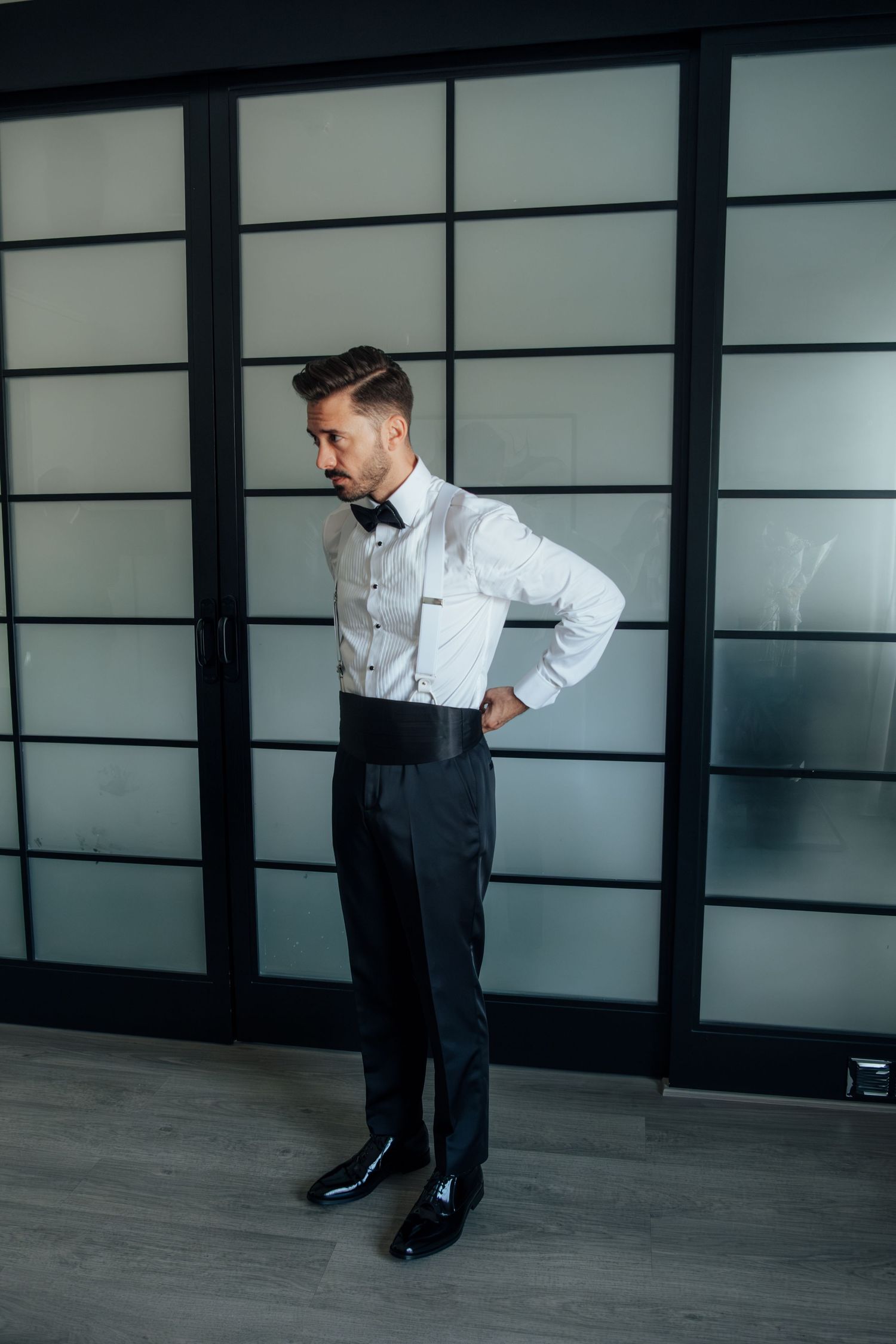 Groom in white shirt, black bow tie, suspenders, and dress pants poses confidently in a modern room.