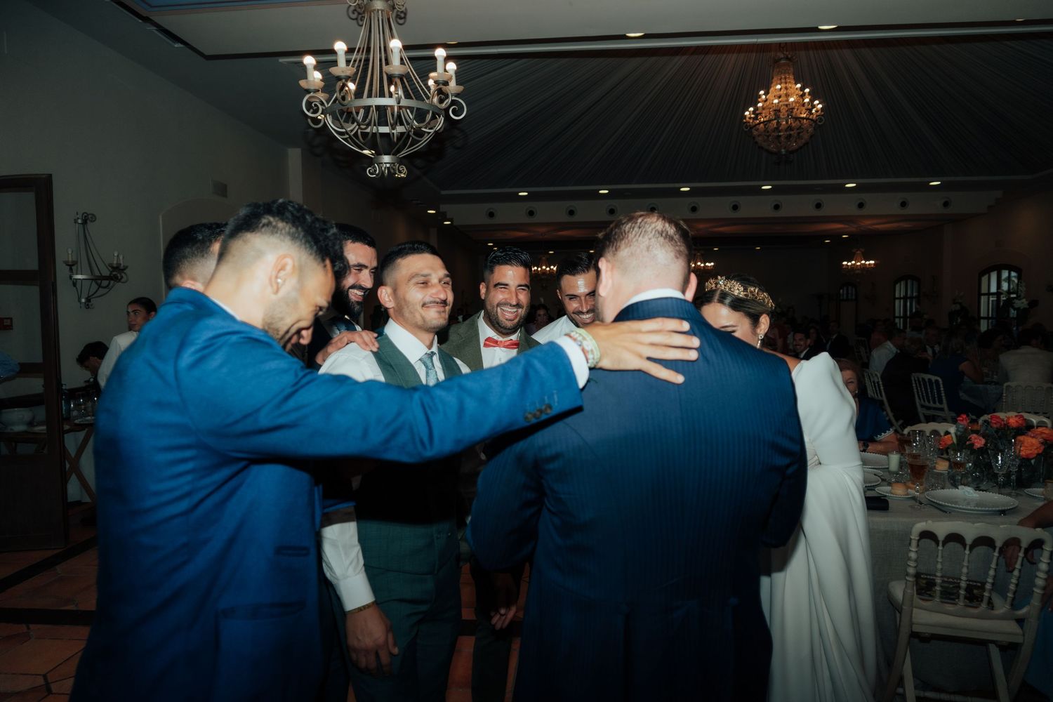 Group of men in suits gather around groom sharing congratulatory embraces at wedding reception.