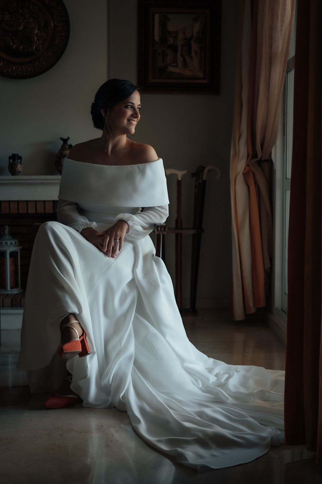 Elegant bride in an off-shoulder white wedding gown sitting gracefully by a window, smiling softly in a dimly lit room.