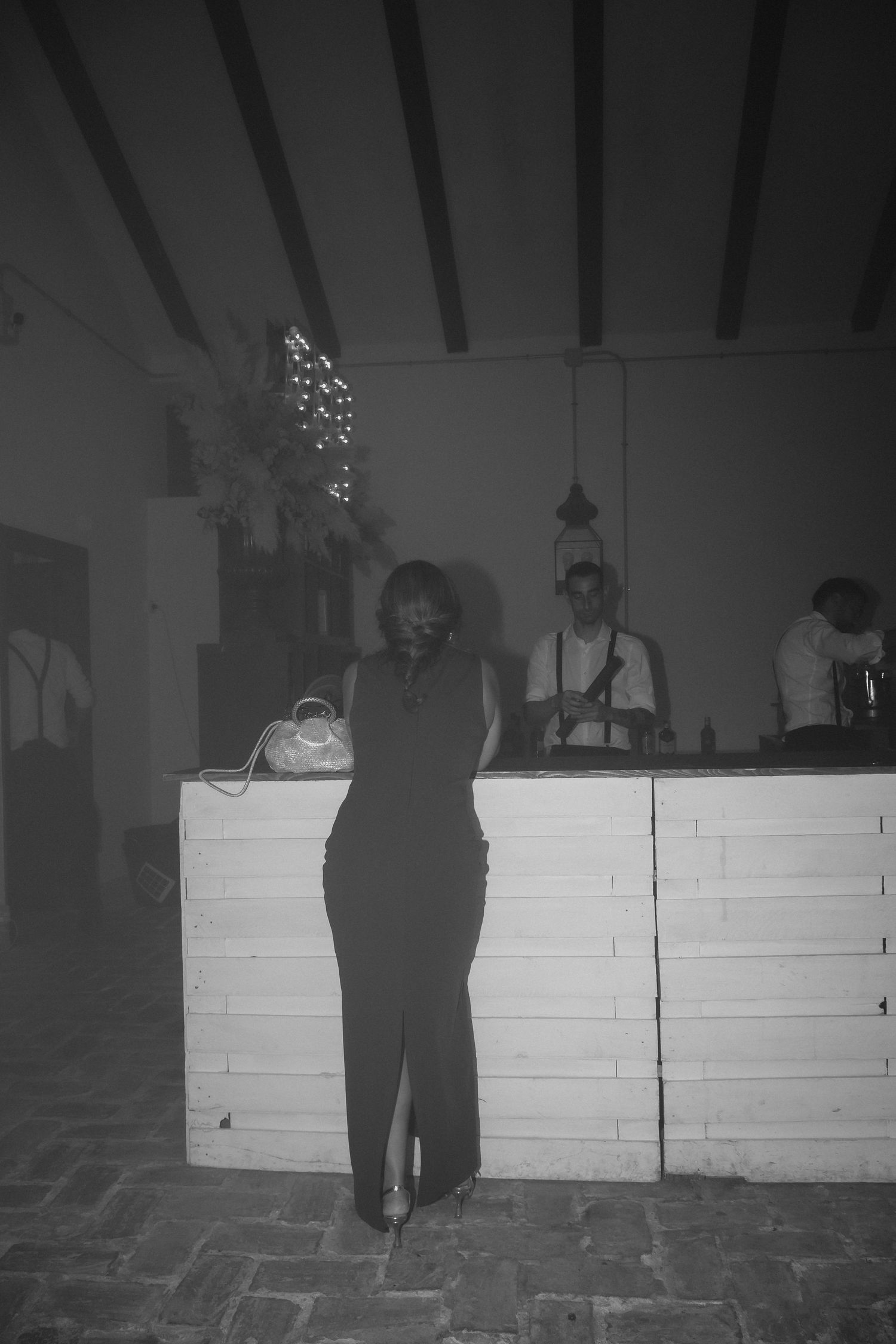 Black and white photo of woman in elegant dress standing by white bar counter at event.