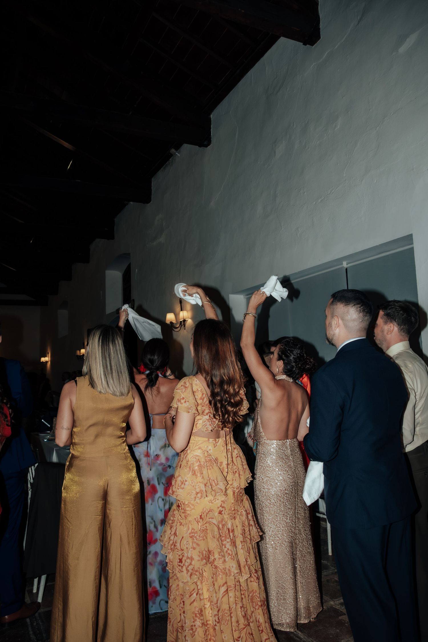 Wedding guests in formal attire waving white napkins in celebration at an indoor evening reception.
