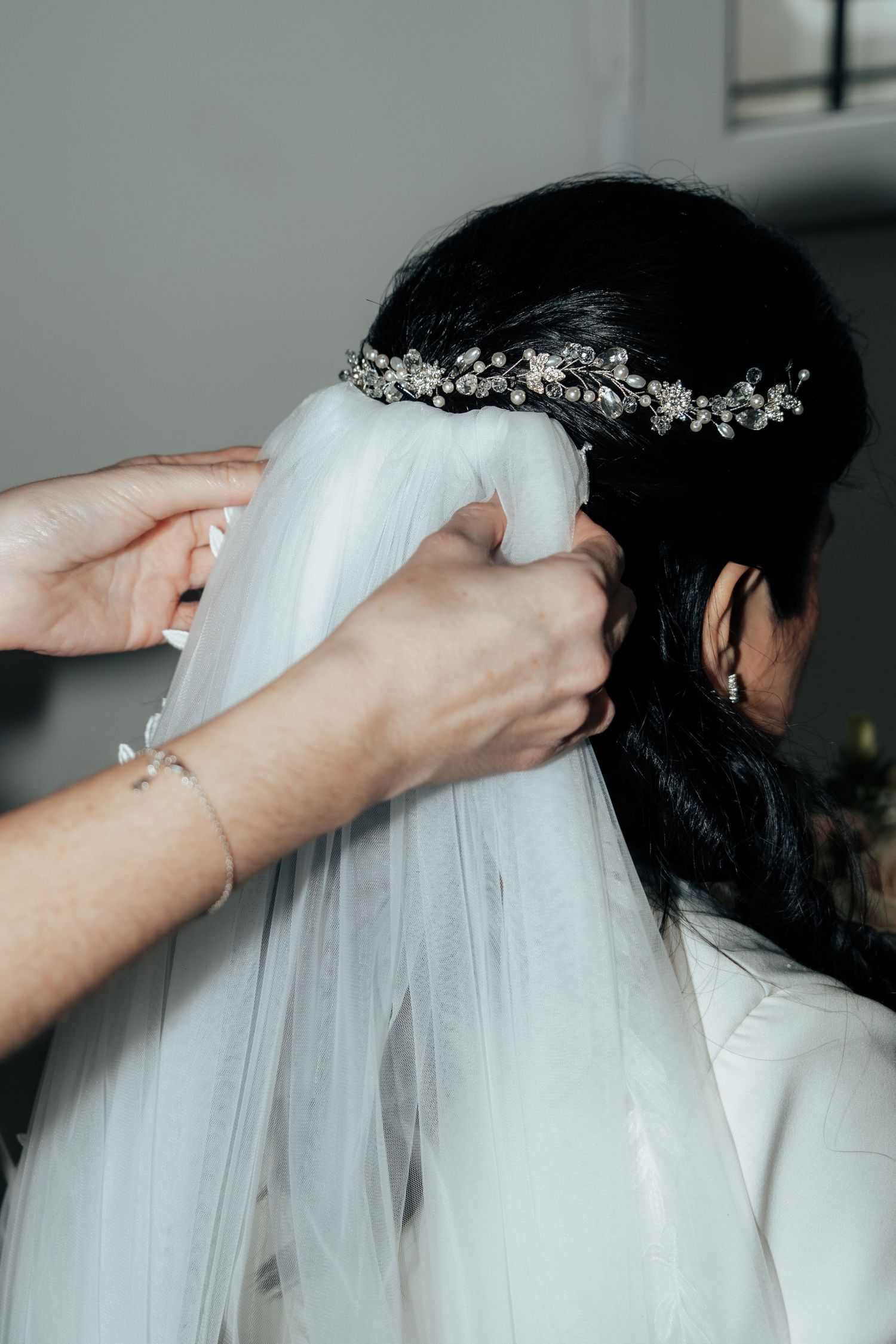 Hands adjusting floral crystal hair vine and white veil on dark-haired bride's updo hairstyle.