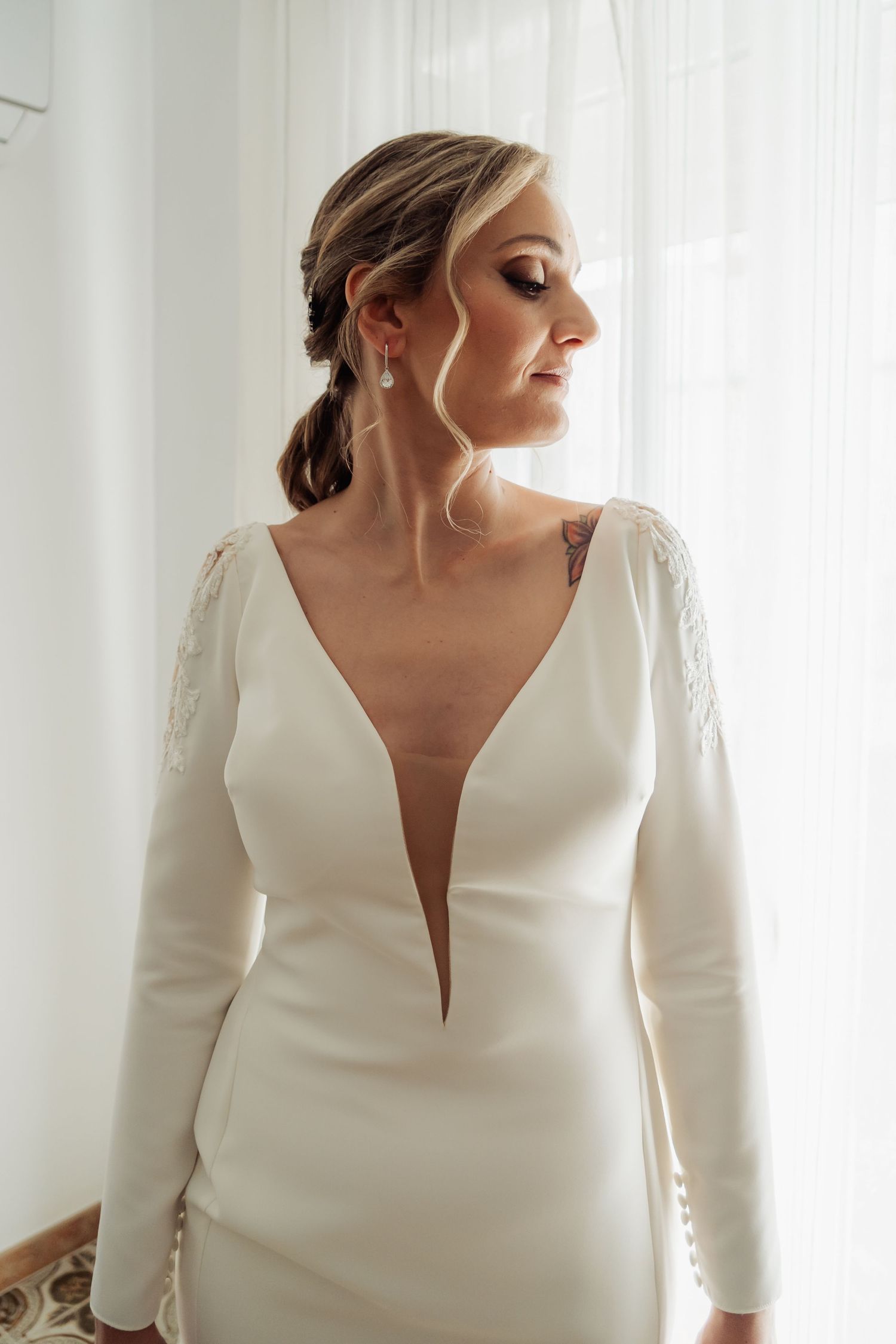 Elegant bride in deep V-neck long-sleeve white gown with pearl earrings standing by window.