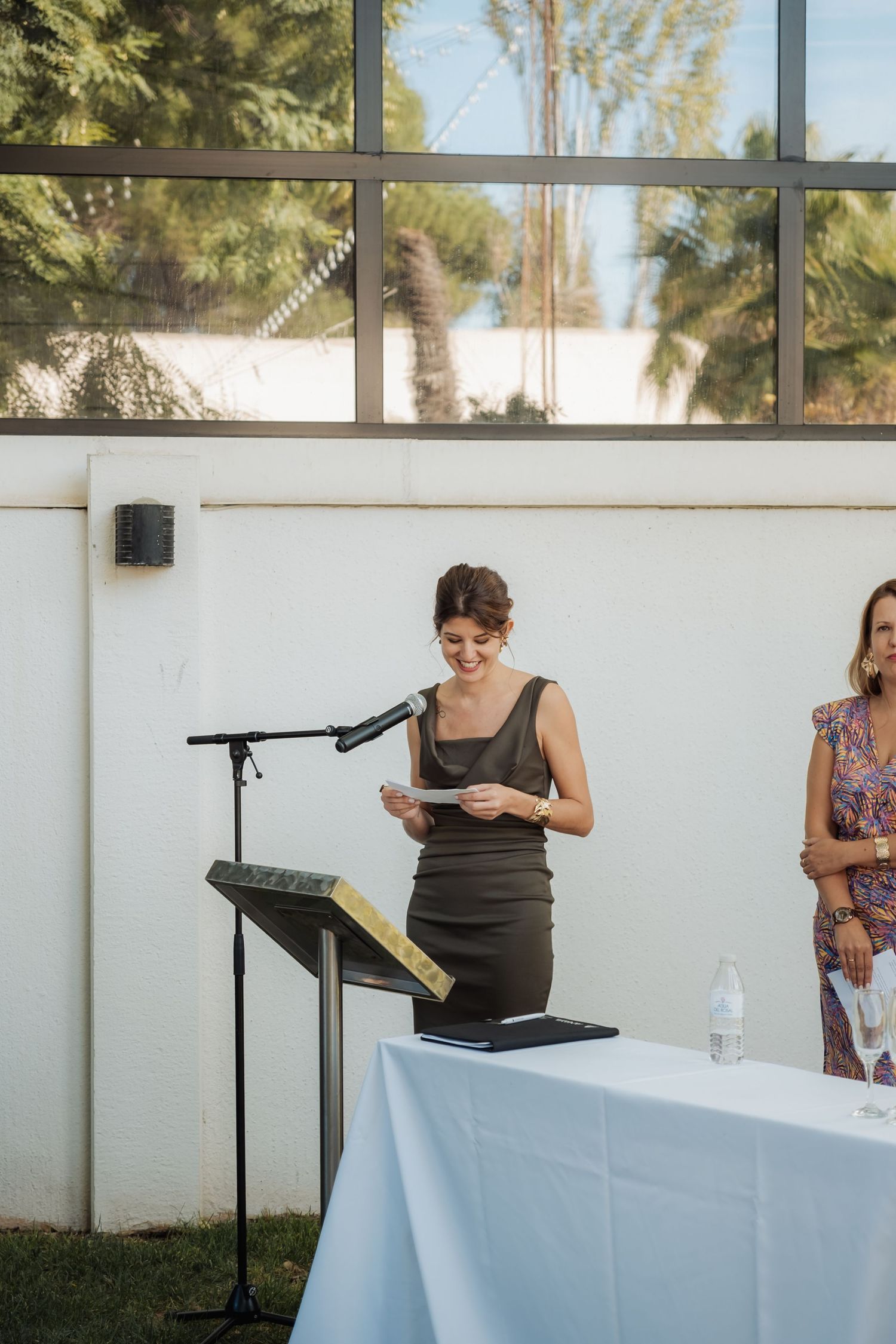 A smiling woman in a grey dress reads from papers at a podium with microphone during an outdoor wedding ceremony.