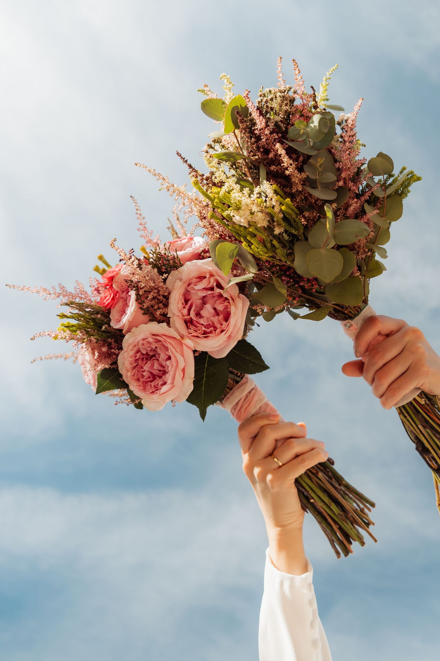 Two hands lift bridal bouquets with pink roses and eucalyptus into a partly cloudy blue sky.