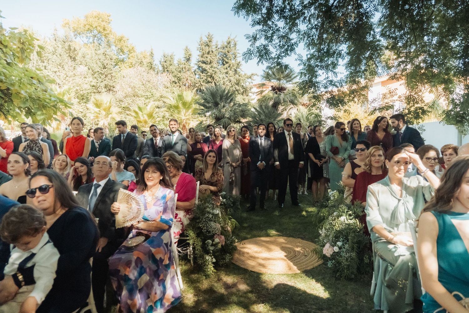 Large group of wedding guests gathered outdoors in garden during sunny ceremony.