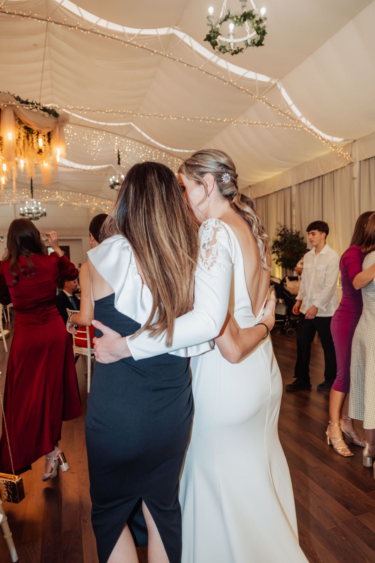 Bride in backless white gown sharing emotional embrace with guest on wedding dance floor.