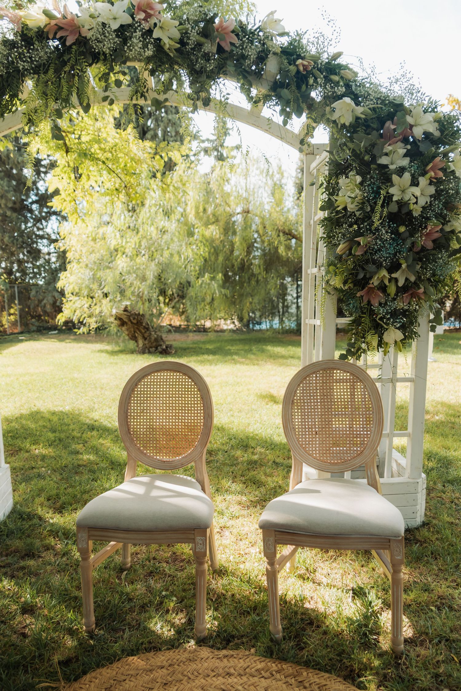 Two elegant rattan chairs sit before a white floral arch adorned with lilies at an outdoor wedding.