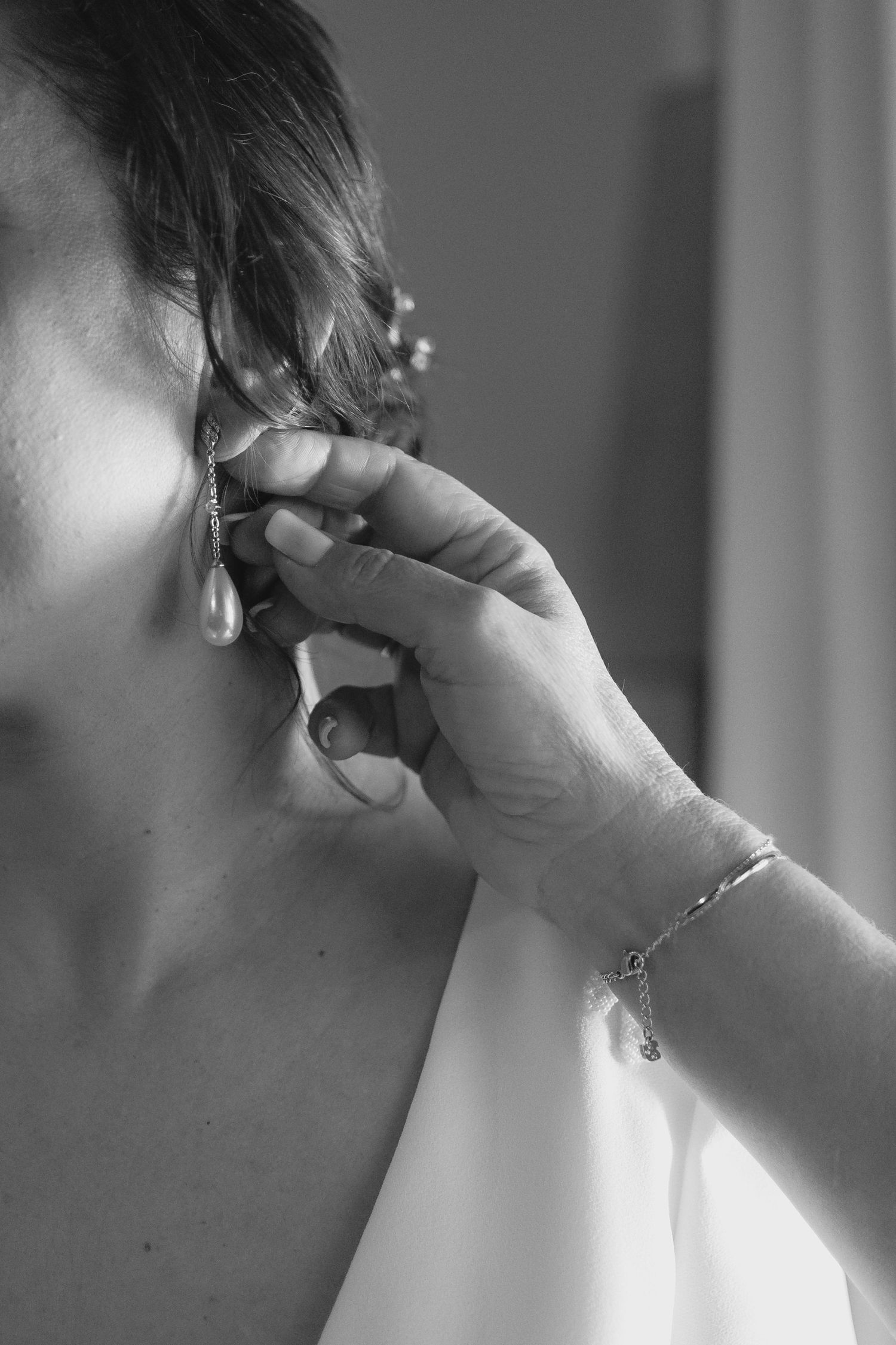 Black and white close-up of hands placing pearl drop earring on bride before wedding ceremony.