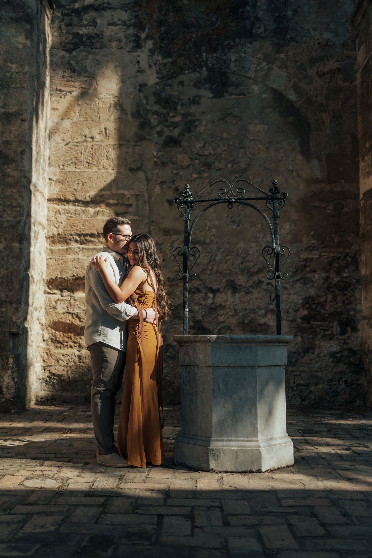 Romantic couple embracing near an ornate stone well against an ancient textured wall in warm sunlight.