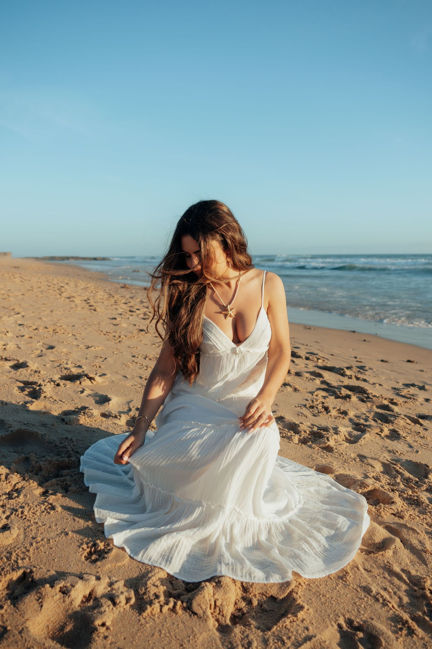 Woman in white flowing dress standing on sandy beach with ocean waves and clear blue sky backdrop.
