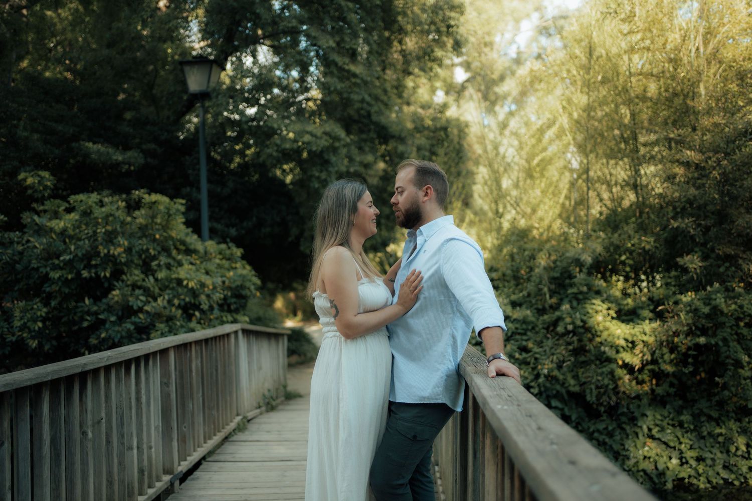 A bride in a white dress and groom in a blue shirt share a tender moment on a sunlit wooden bridge.