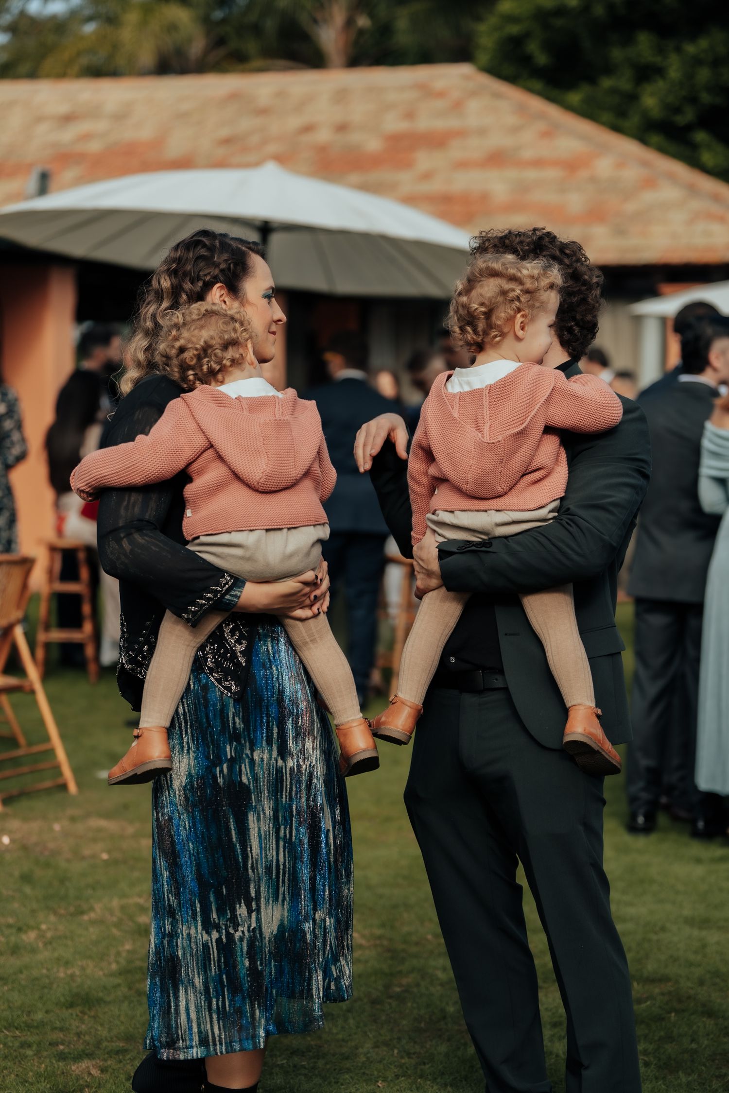 Parents carry twin toddlers in matching pink hoodies at an outdoor wedding reception on green grass.
