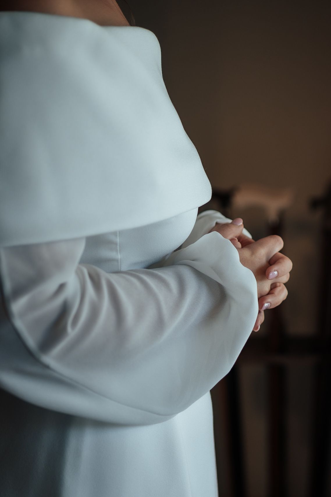 Close-up of a bride in an elegant off-shoulder white wedding dress with her hands clasped together.