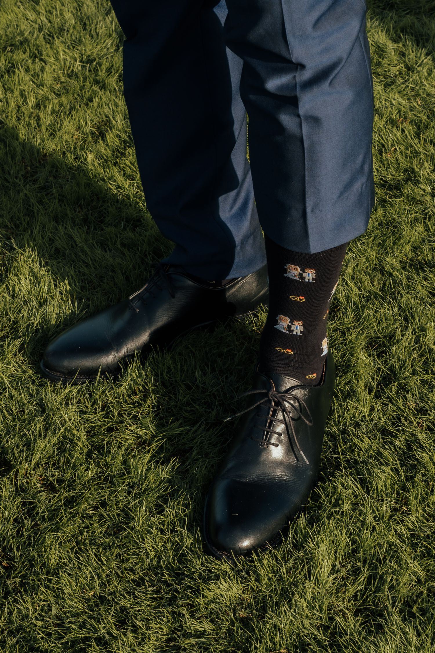 Groom wearing patterned socks and black dress shoes standing on green grass.