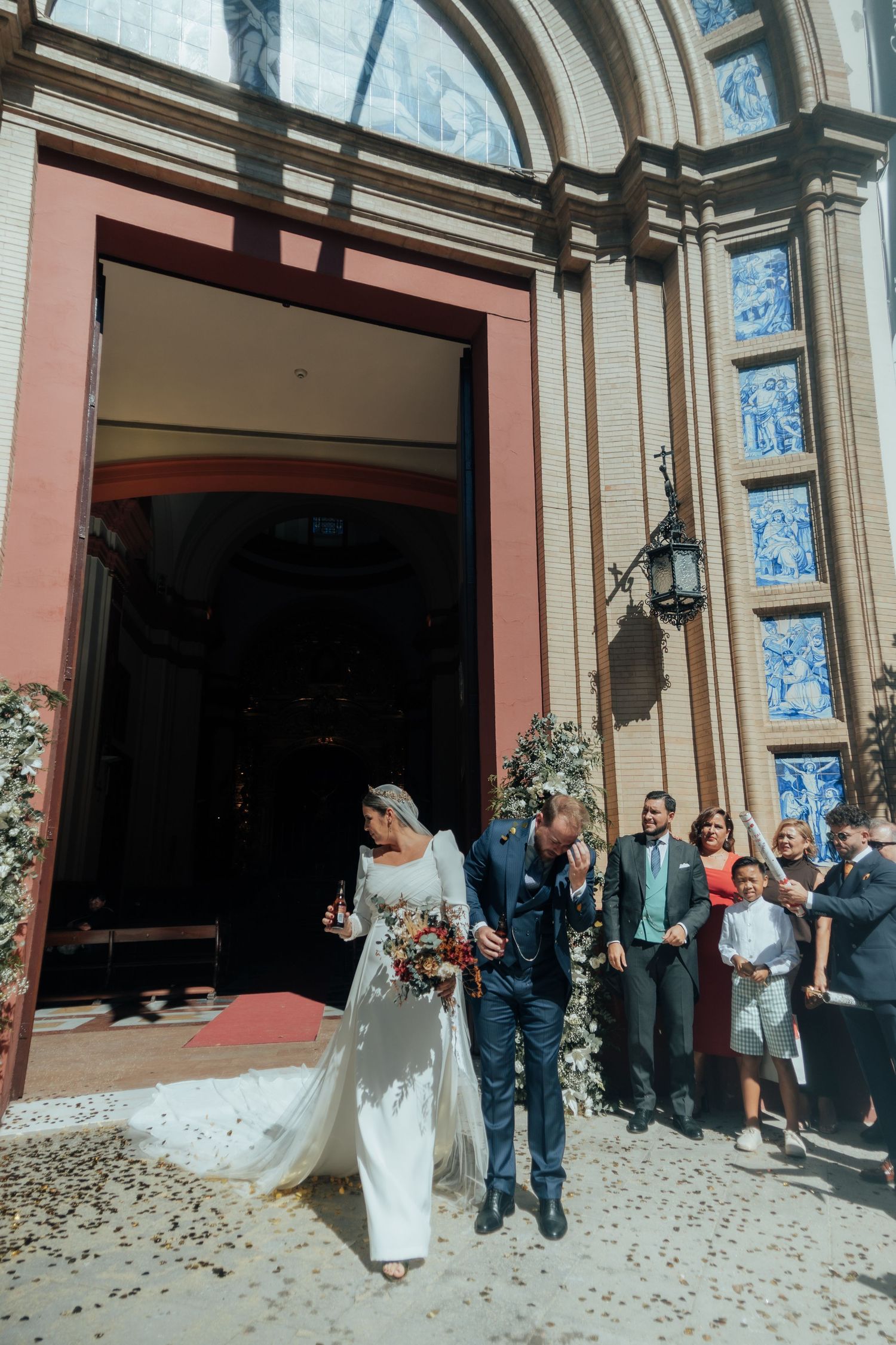 Bride in white gown with bouquet and groom exit ornate church with blue tile facade.