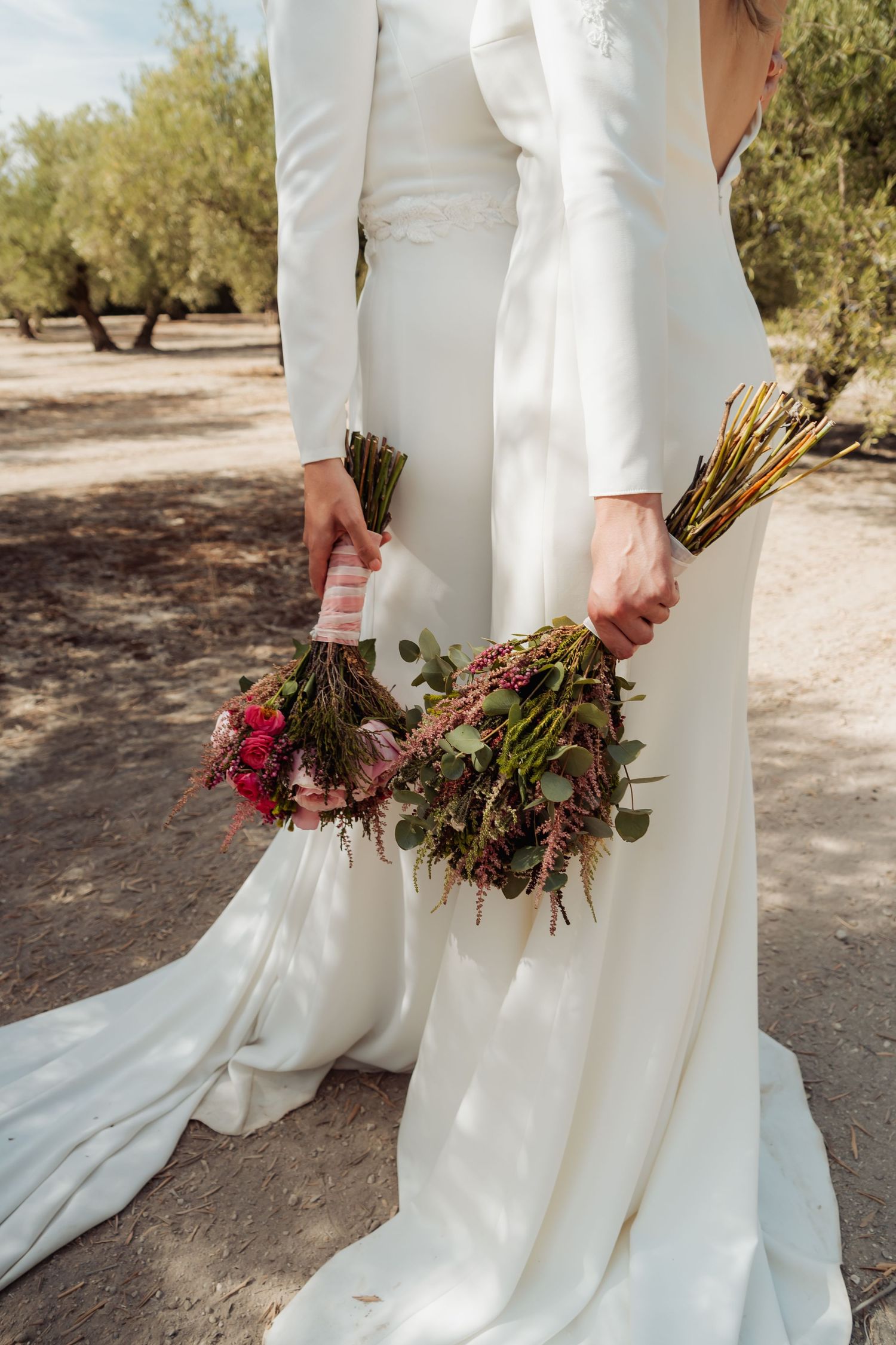 Two brides standing back to back holding colorful wildflower bouquets in an olive grove.