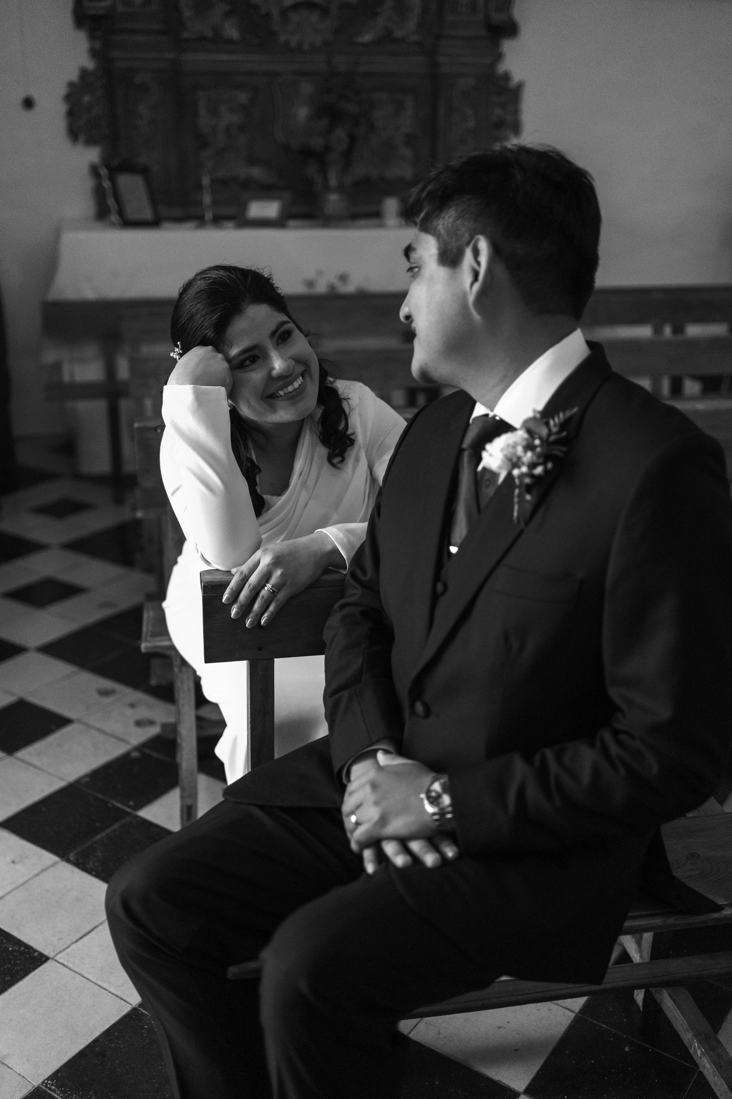 Black and white photo of a smiling bride and groom sitting together in a church before their wedding ceremony.