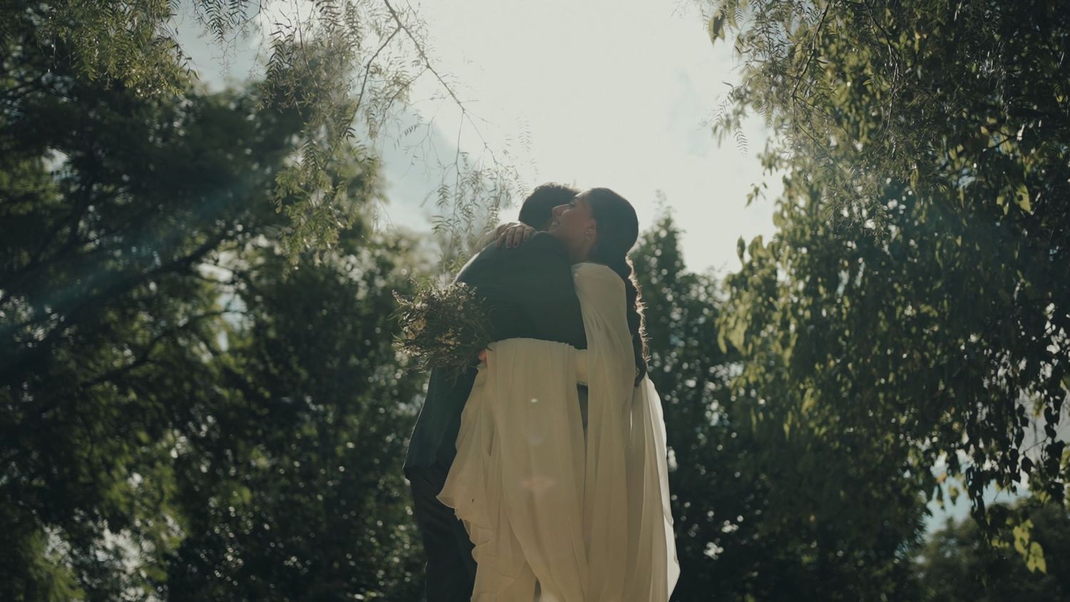 A wedding couple shares a romantic kiss surrounded by sunlit trees in an outdoor setting.
