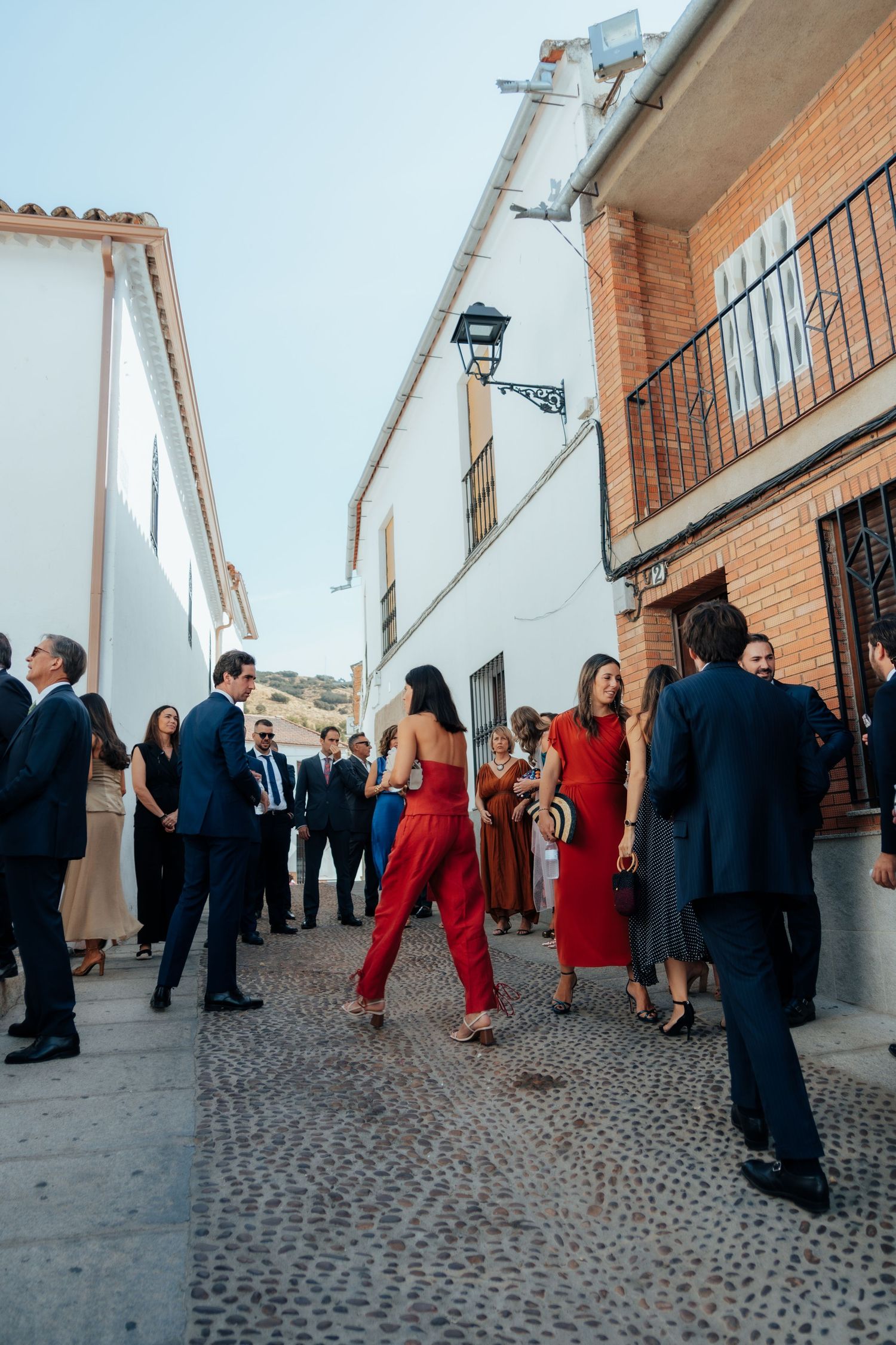 Wedding guests in formal attire gathering on cobblestone street outside Spanish white-washed buildings.