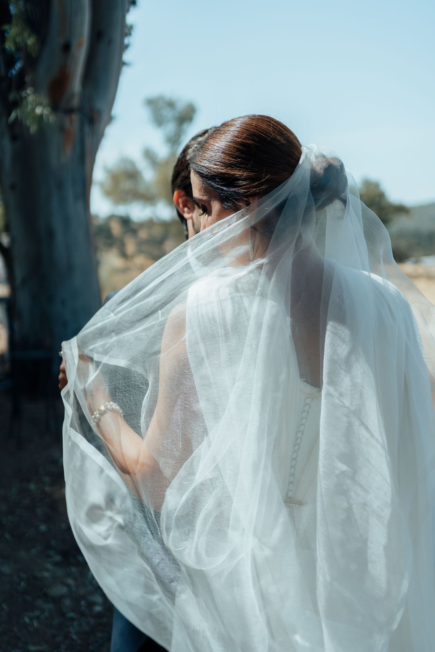 Bride in white wedding dress with veil draped over her shoulders standing outdoors in bright sunlight.