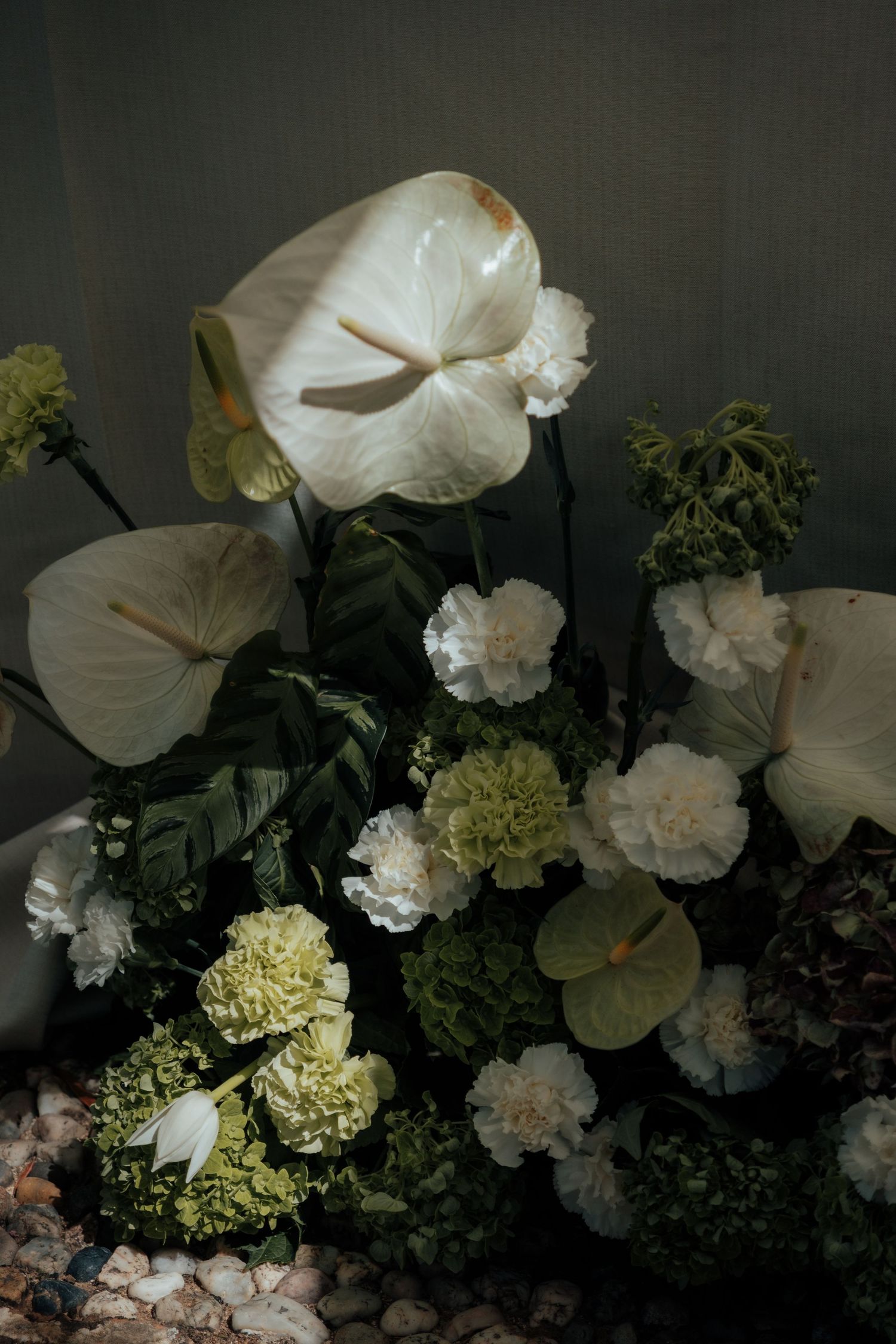Elegant floral arrangement featuring white anthuriums, carnations, and green hydrangeas on a bed of pebbles.