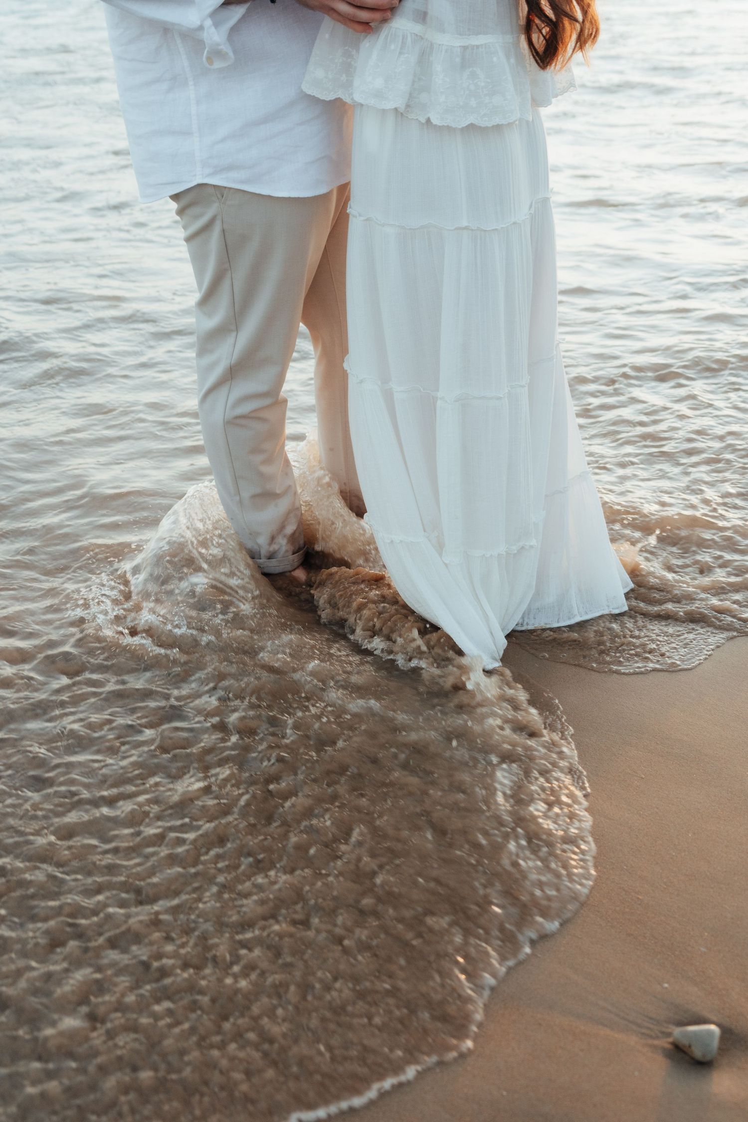 Couple standing barefoot on sandy beach in white outfits, waves washing over their feet at sunset.