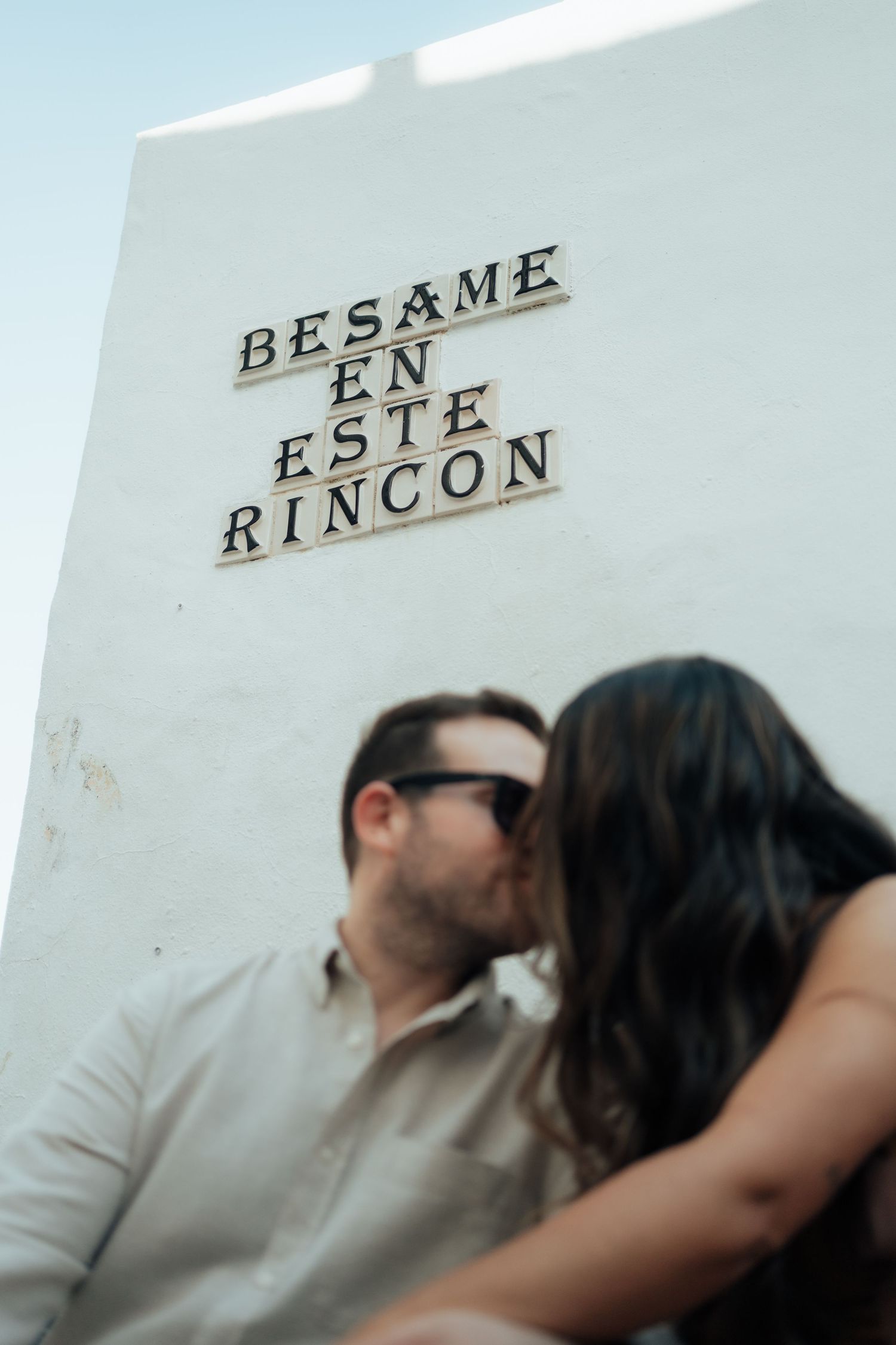 Couple kissing beneath a white wall sign reading Besame En Este Rincon, meaning kiss me in this corner.