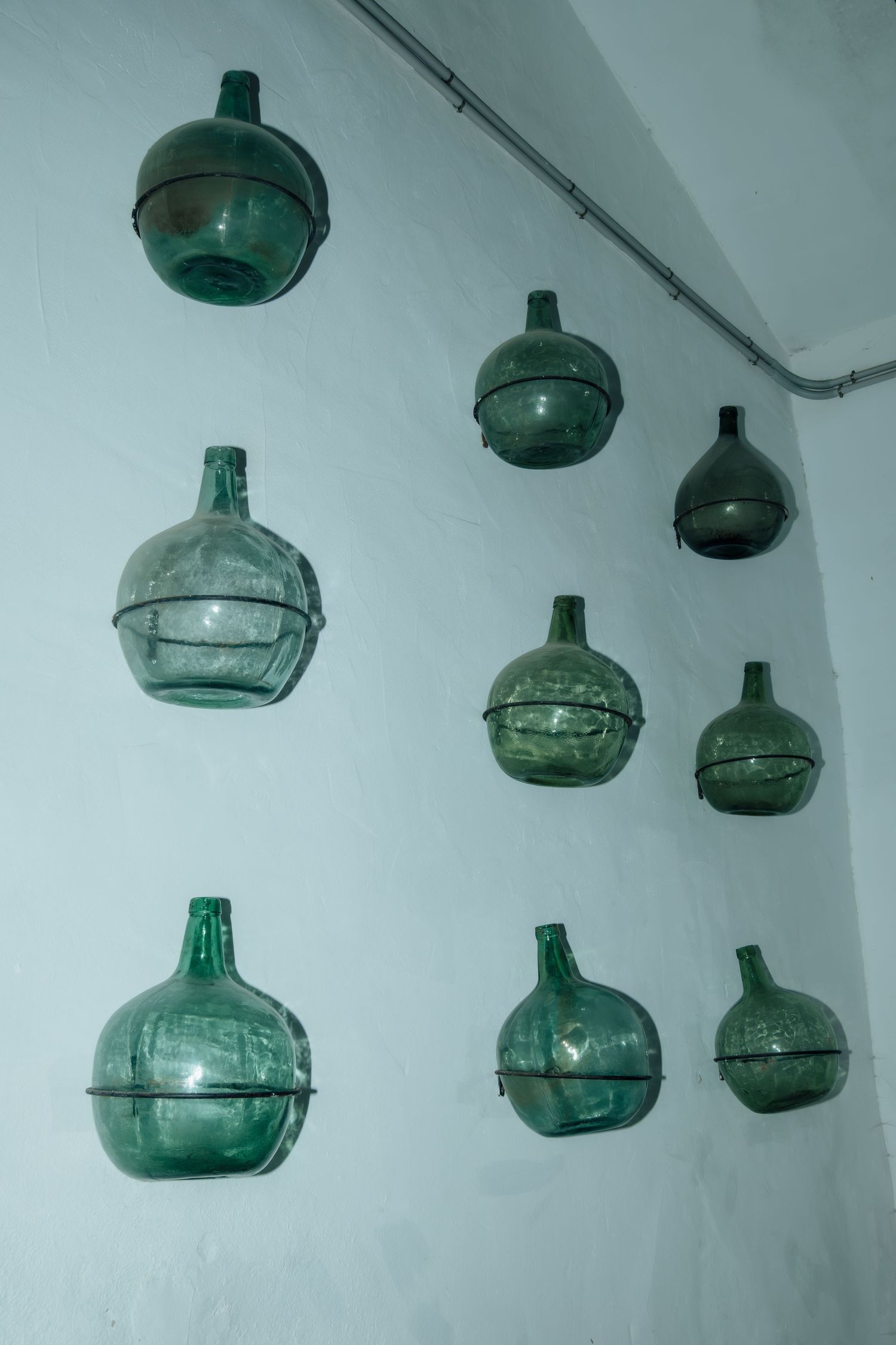 Green glass demijohn bottles mounted decoratively on white wall in rustic wine cellar.