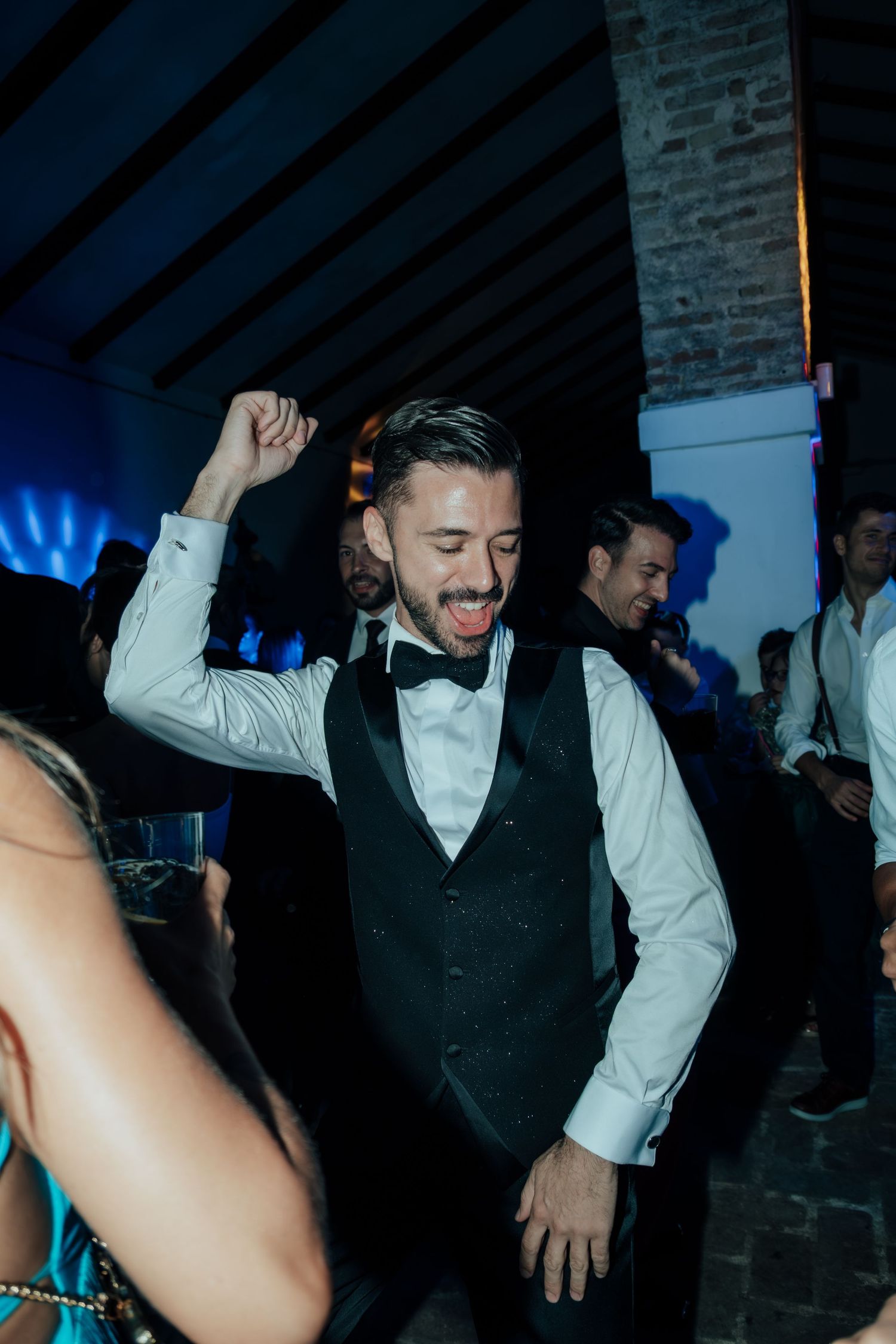 Excited groom in vest and bow tie dancing with fist raised at wedding reception party.