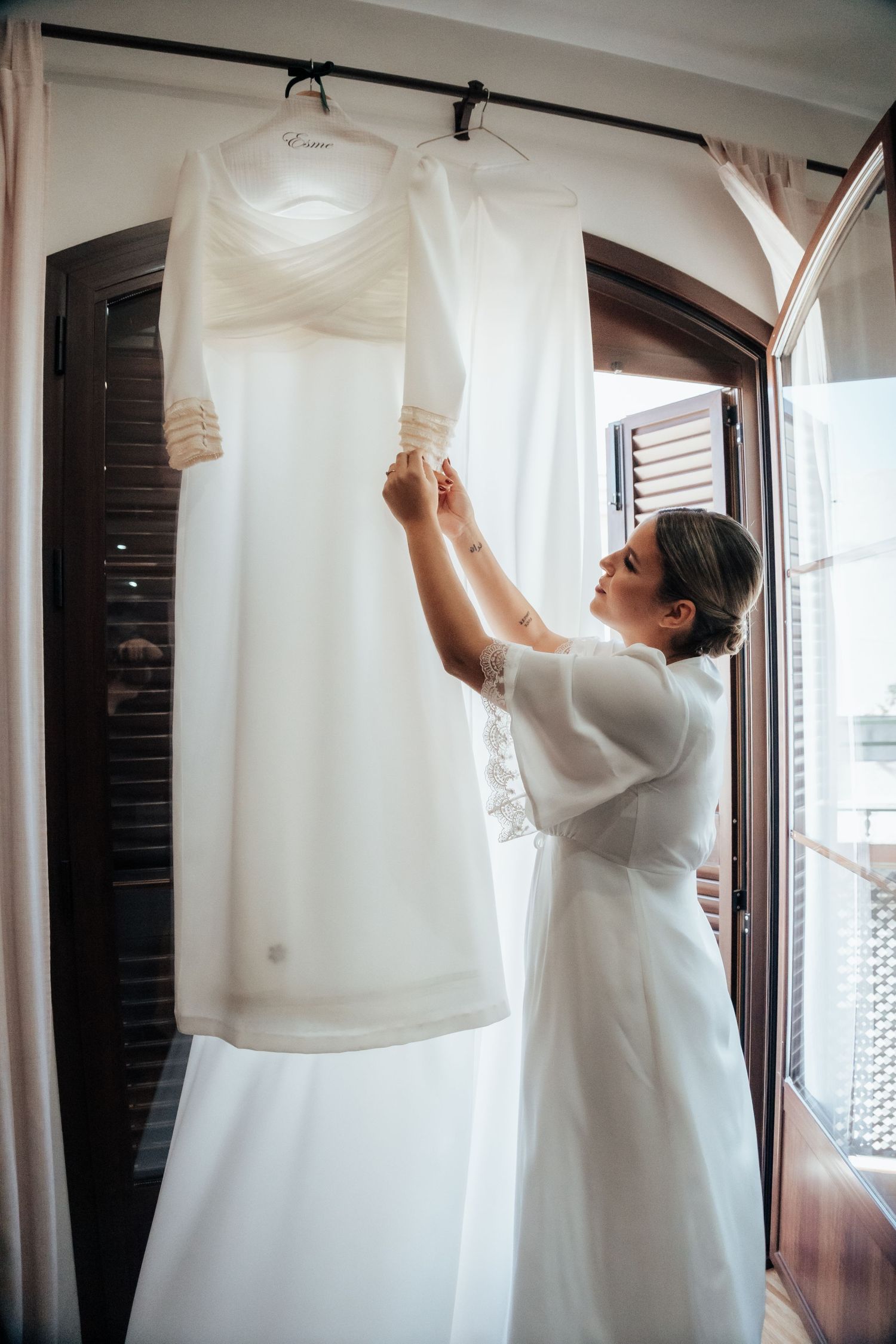 Bride in white robe admiring her wedding dress hanging near an arched balcony door.