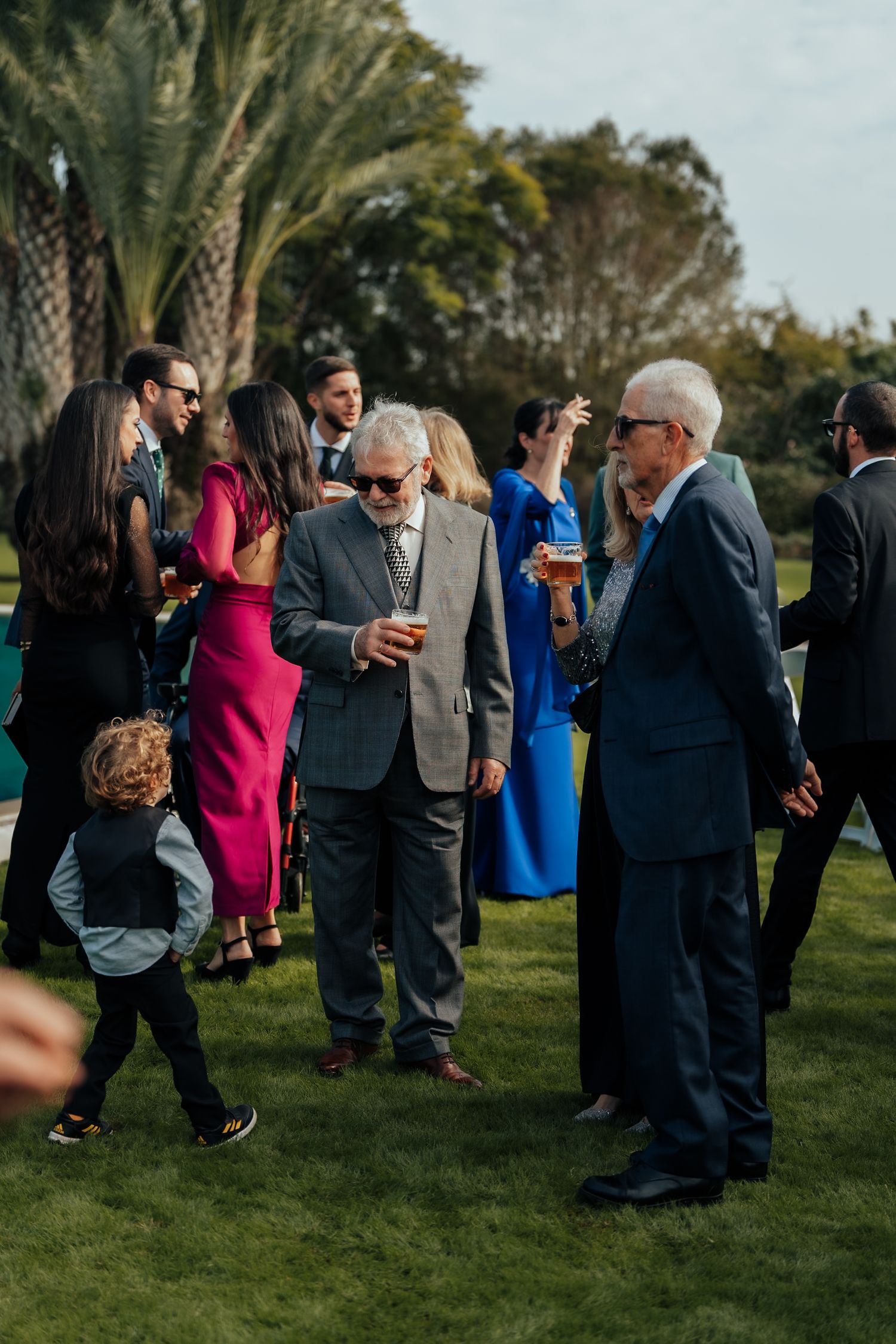 Elegantly dressed wedding guests mingle on lush green lawn, with men in suits and women in colorful formal dresses.