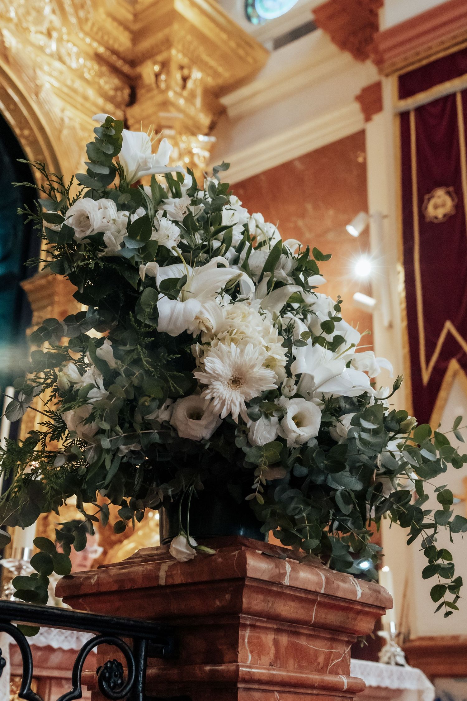 Elegant white floral arrangement with roses and eucalyptus on marble pedestal in ornate church.