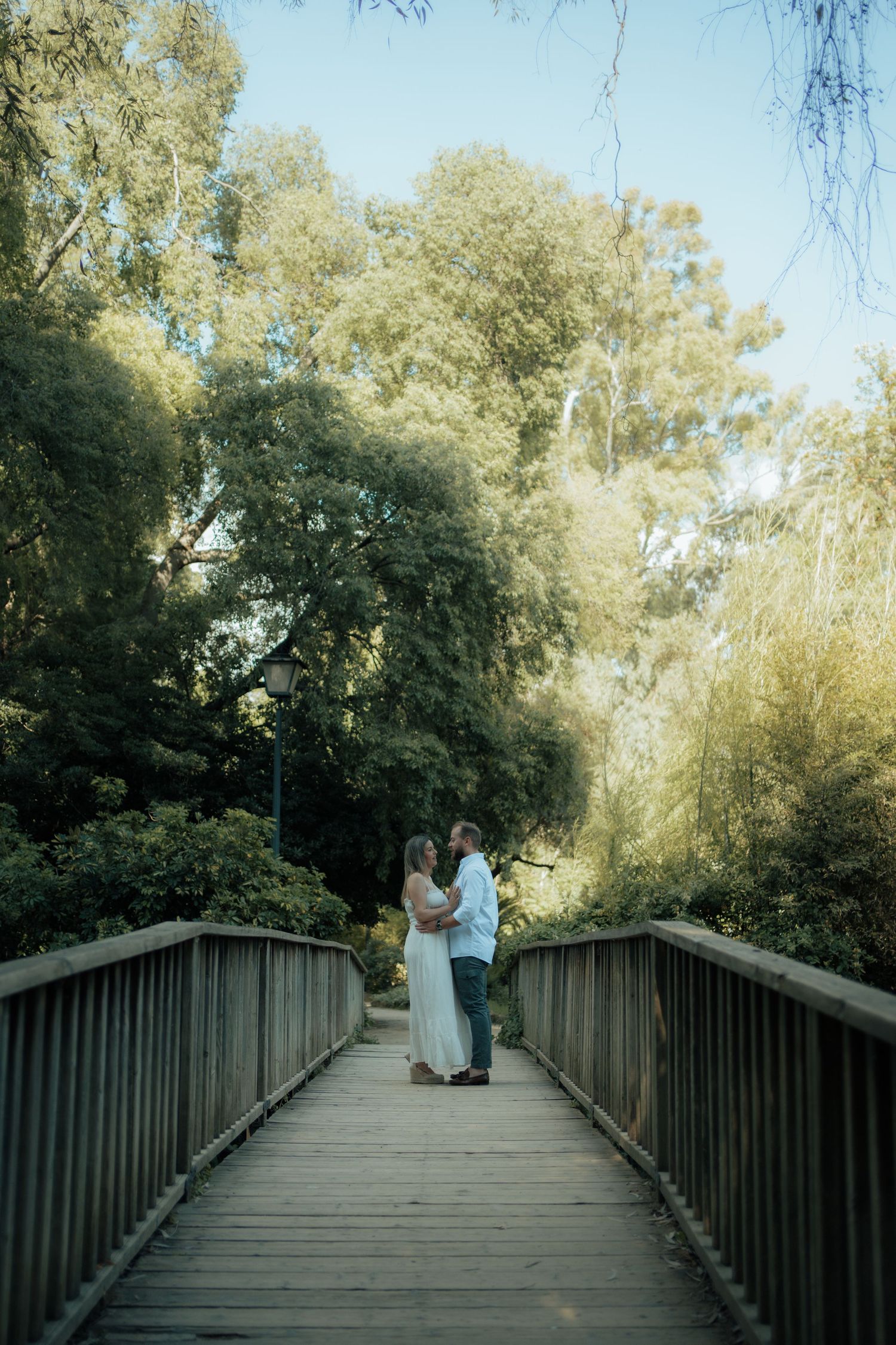 A couple in wedding attire share a romantic moment on a wooden bridge surrounded by lush green trees.