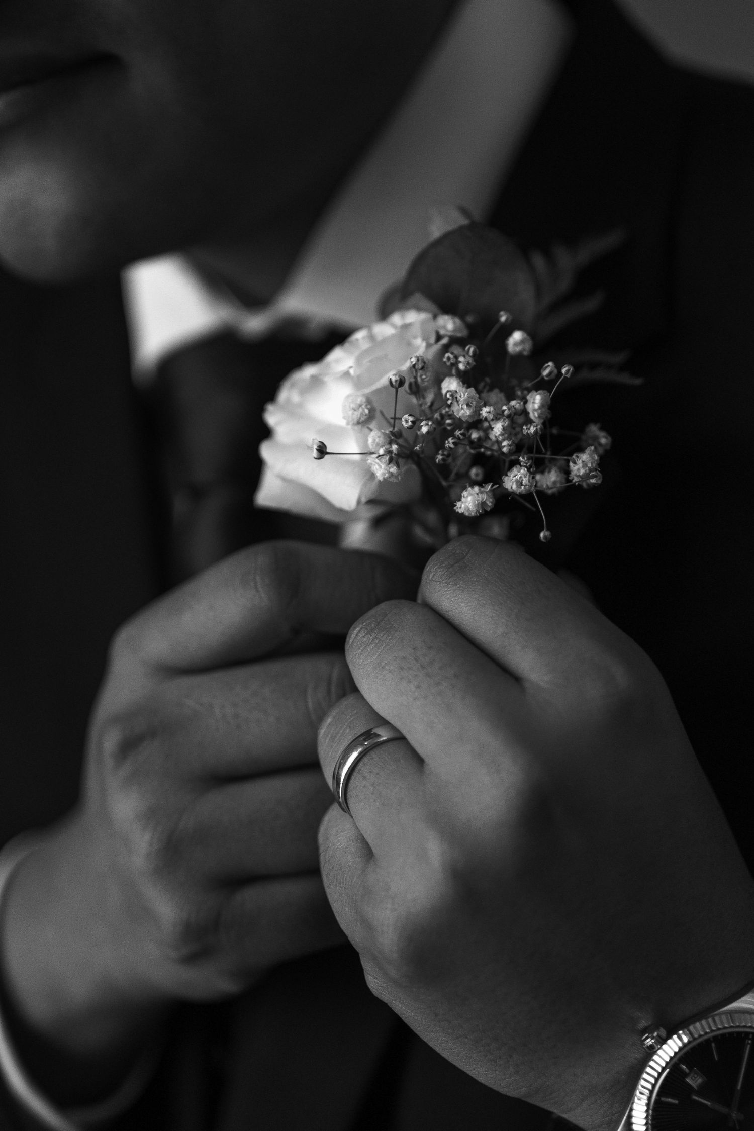Close-up black and white photo of hands pinning a floral boutonniere on a groom's suit lapel.