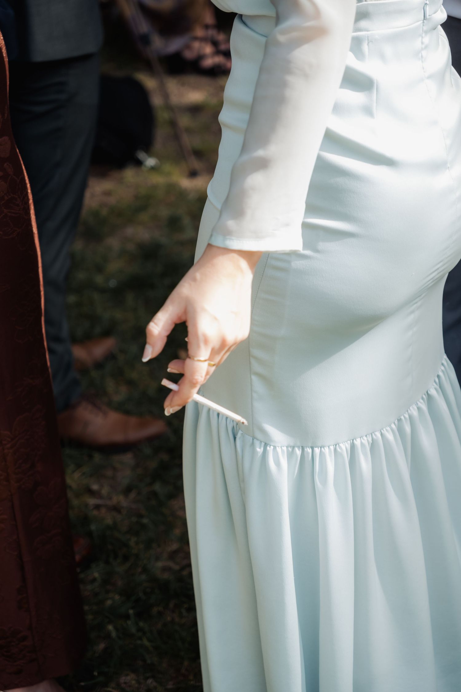 Close-up of a bride in a light blue satin gown holding a cigarette at an outdoor wedding.