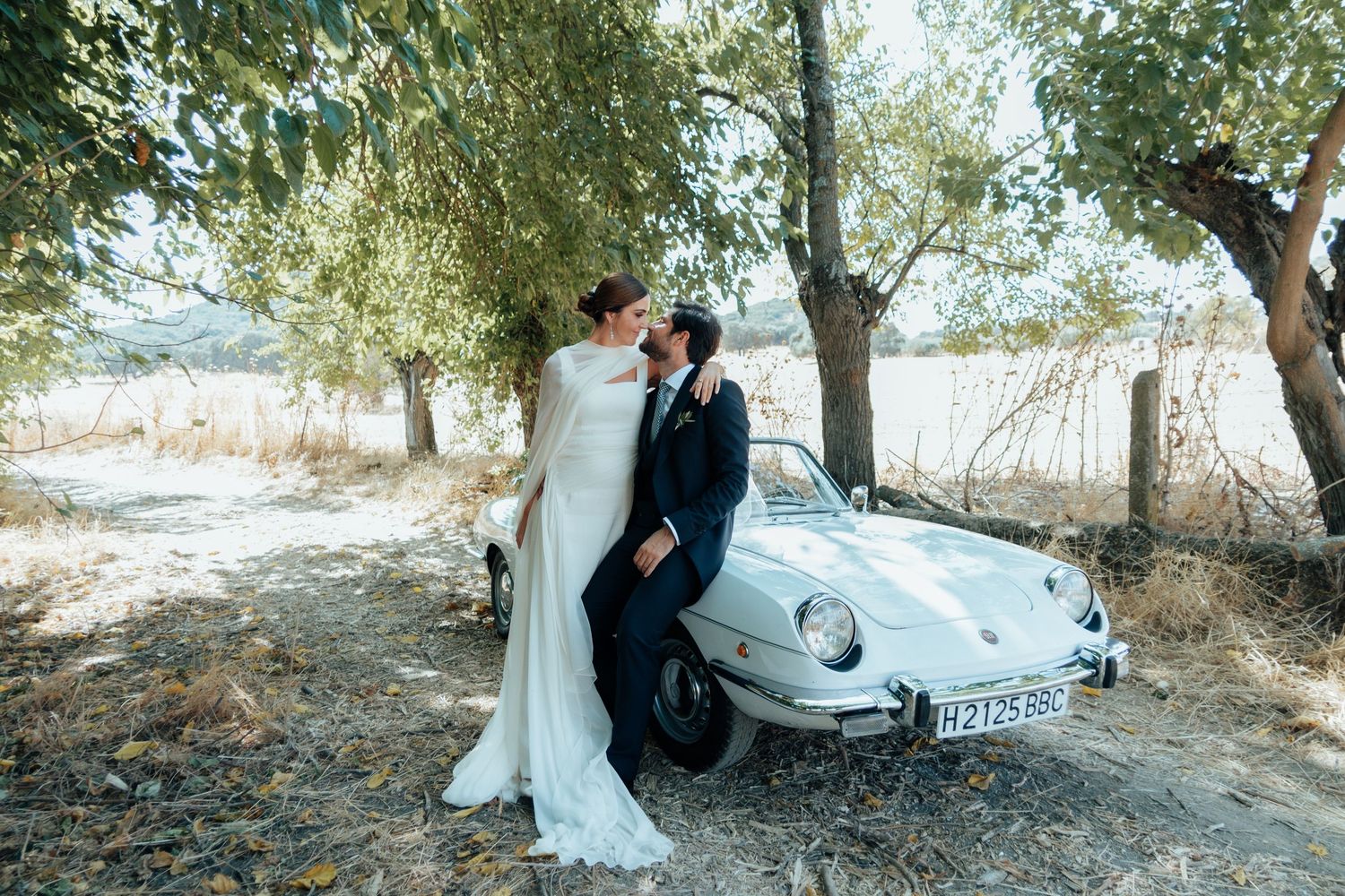 Bride and groom share a romantic kiss beside a vintage white Porsche under shady trees.